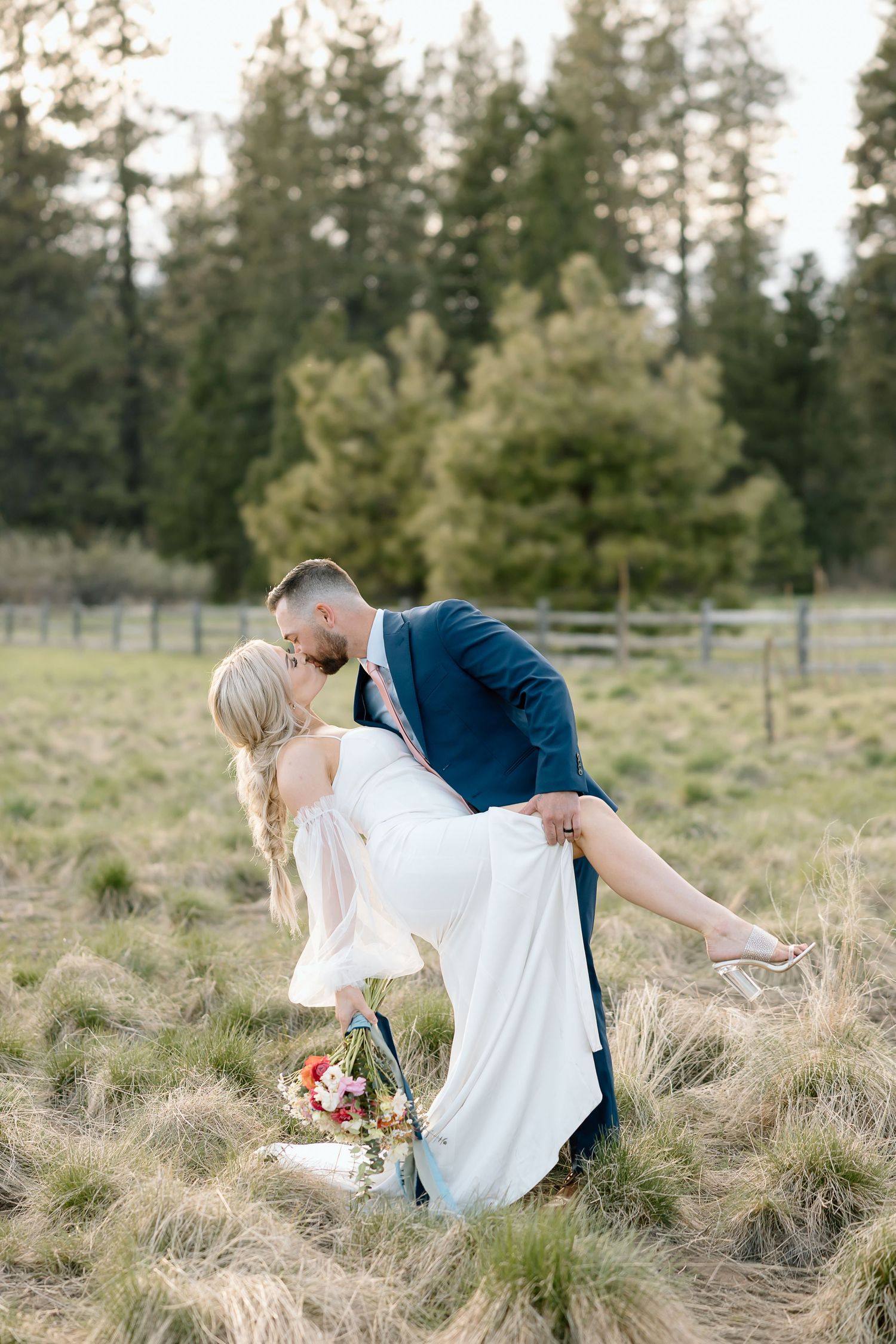 Romantic dip pose of wedding couple in field with bouquet and evergreen forest backdrop.