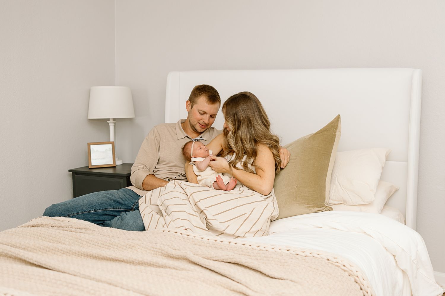 Parents sharing an intimate moment with their newborn while sitting on a cream-colored bed.