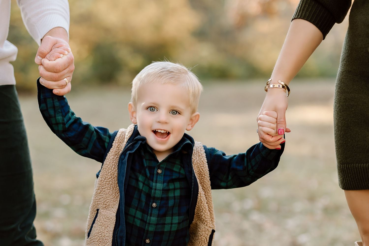 Young child in plaid shirt and vest holding hands with adults in an autumn setting.