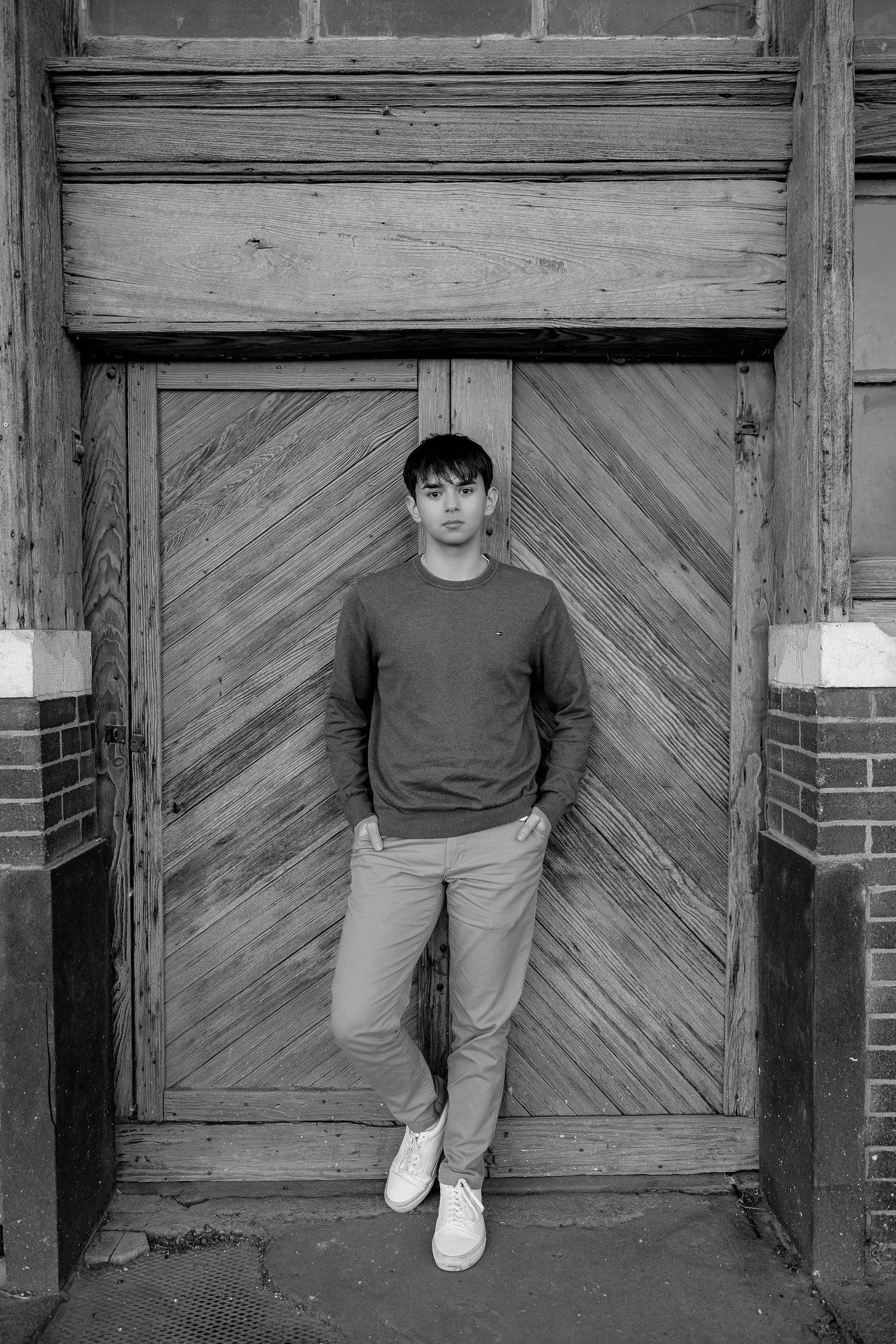 Black and white portrait of person standing against chevron wooden door with brick accents.