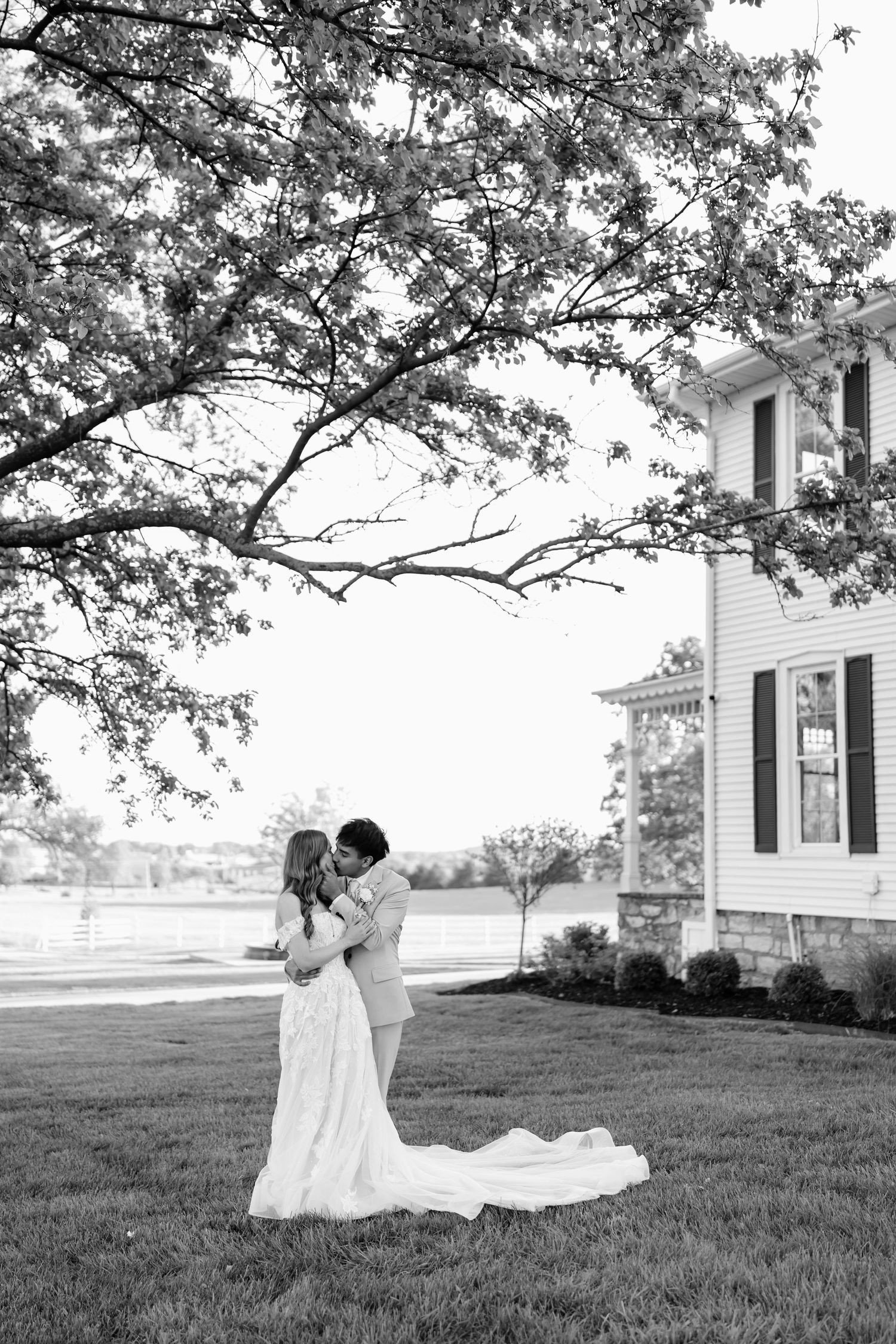 Black and white photo of a bride and groom embracing on a lawn beneath a large tree beside a classic white farmhouse.
