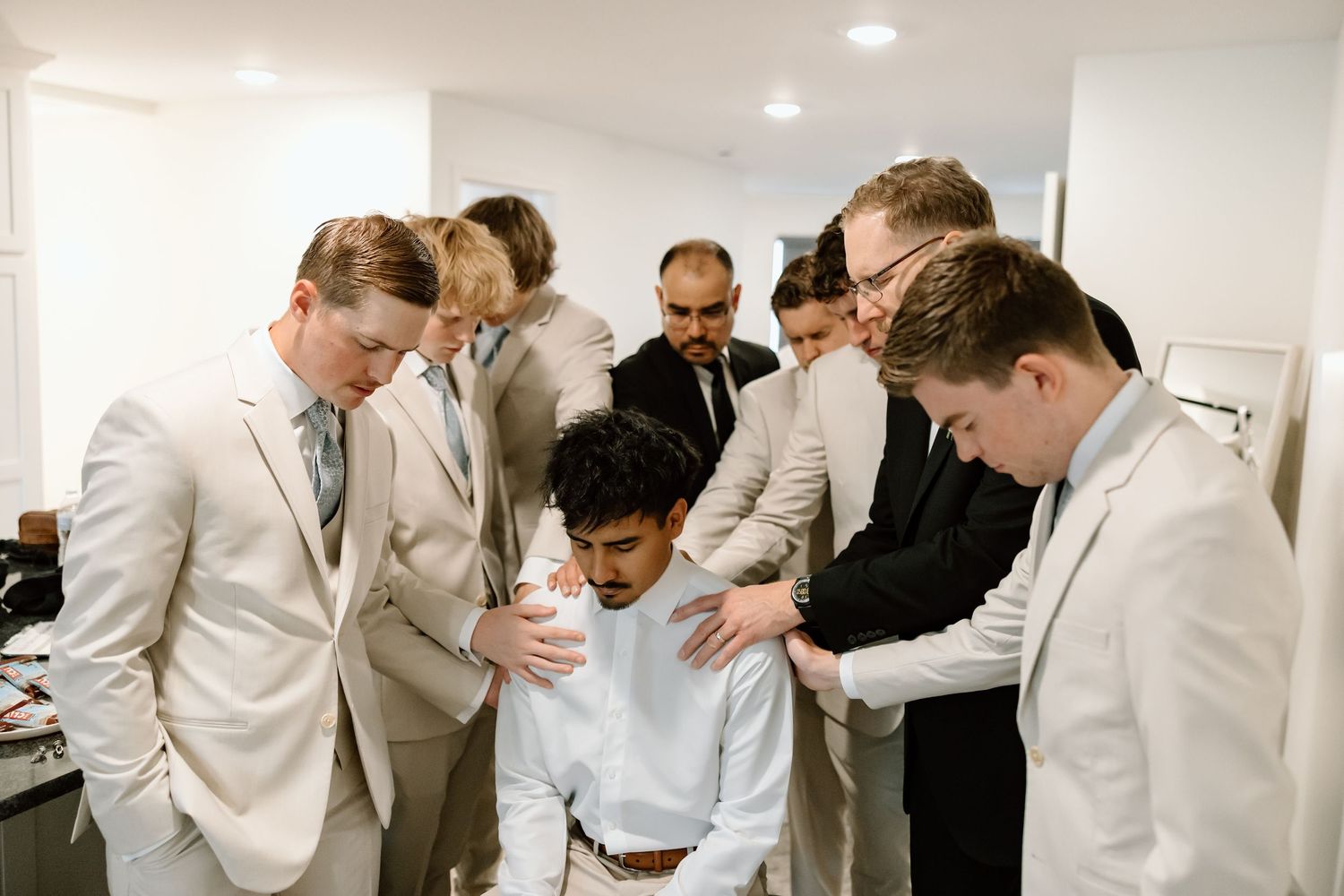 Groomsmen in light suits gather around groom in prayer circle before wedding ceremony indoors.