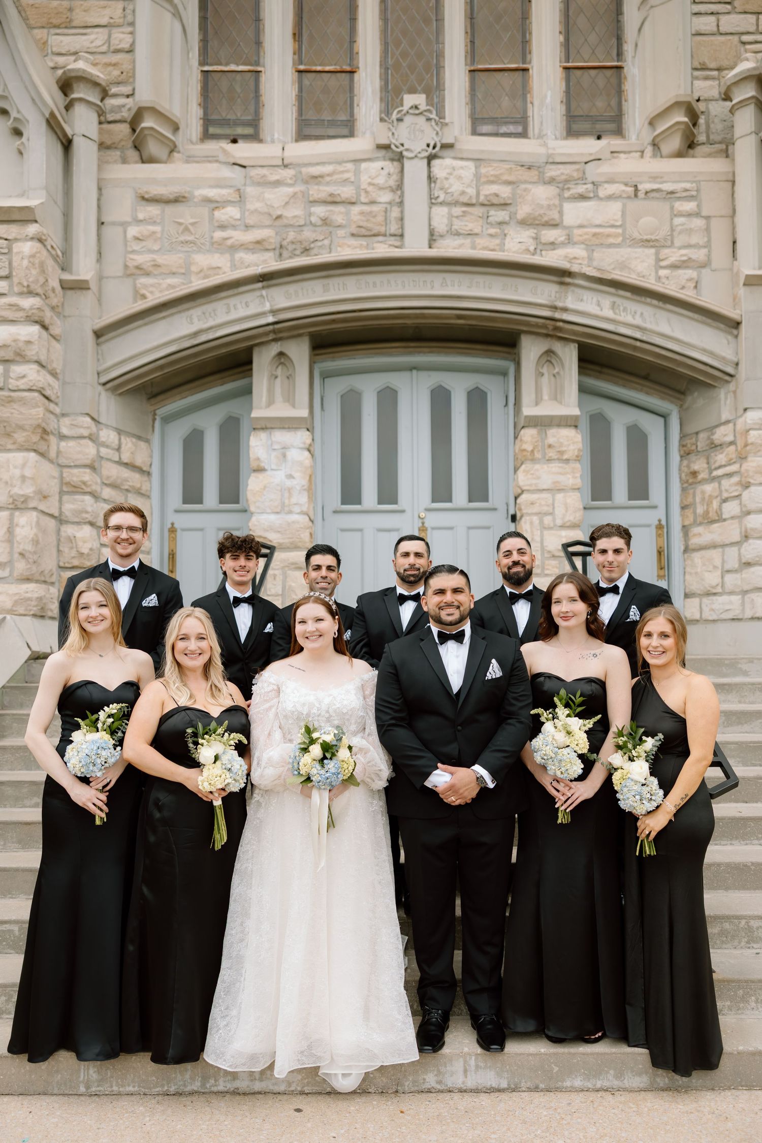 Wedding party poses on stone church steps, bride in white gown with groomsmen in tuxedos and bridesmaids in black dresses.