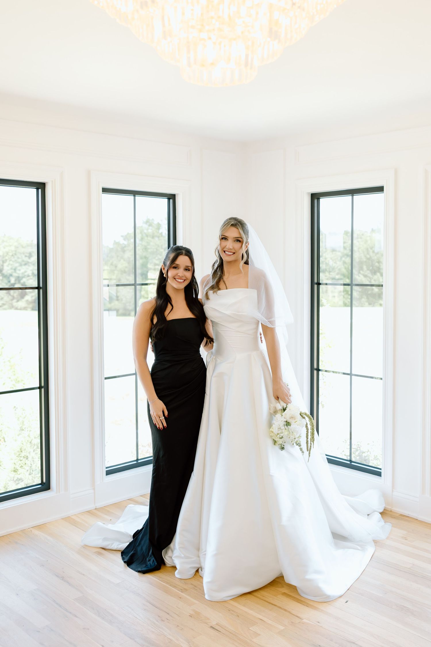 Bride in white gown with veil holds bouquet beside bridesmaid in black dress, posing in bright white room.