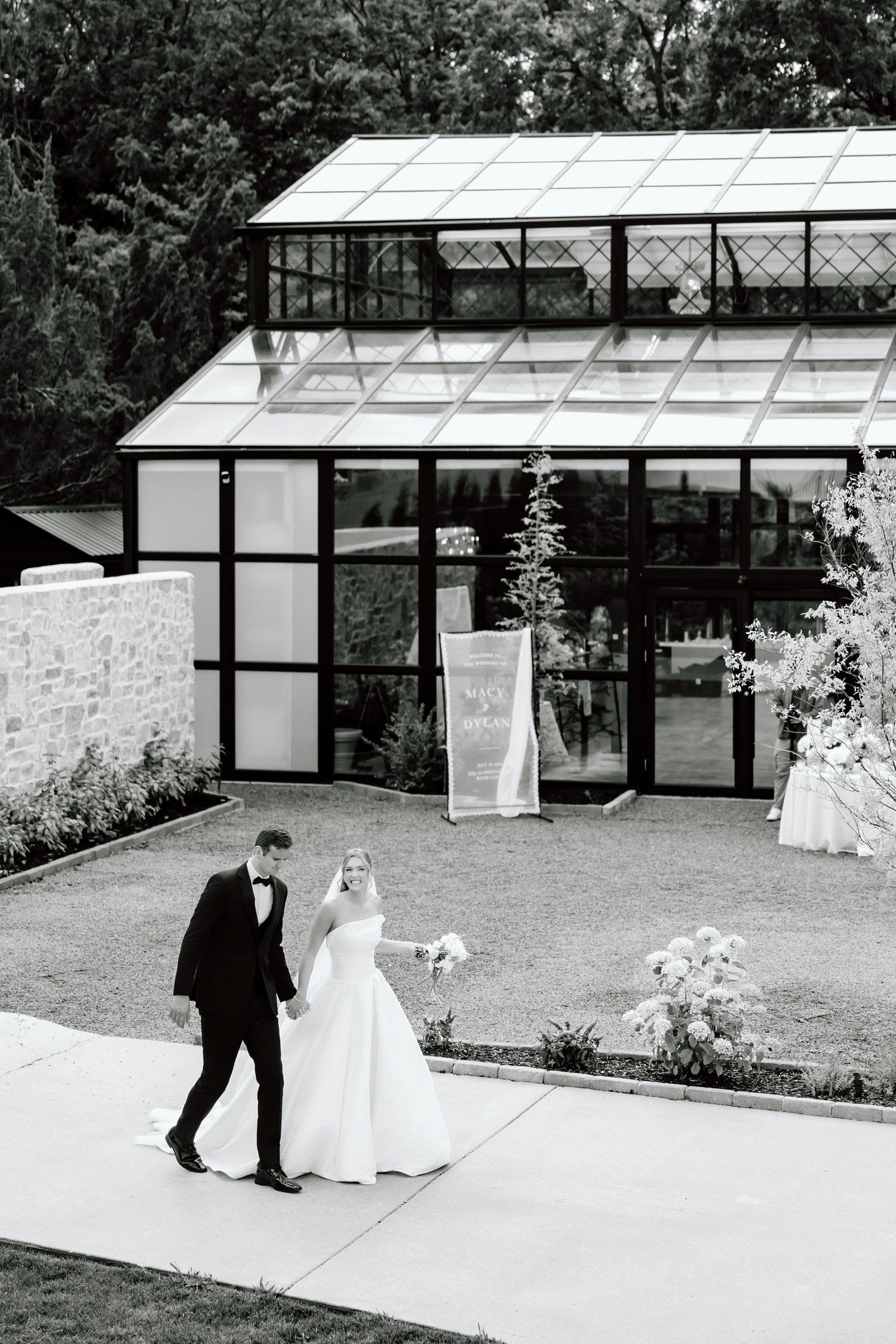 Black and white photo of bride and groom walking hand in hand outside a glass greenhouse wedding venue.