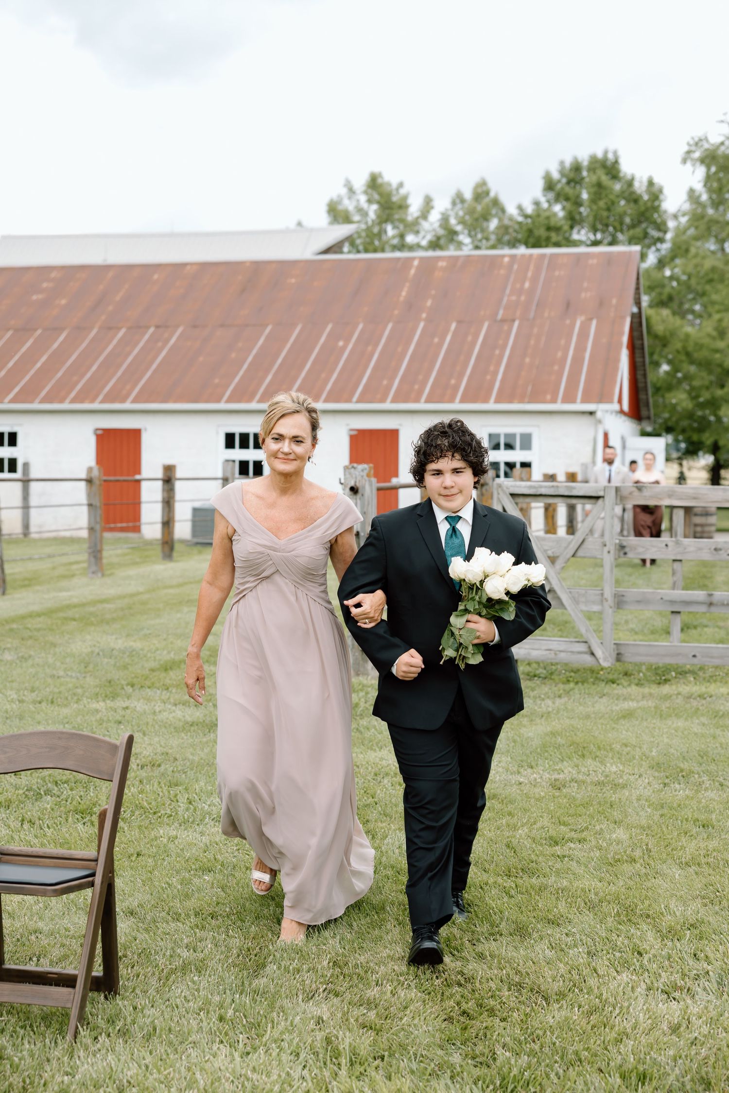 A woman in a taupe gown and a boy in a suit walk arm-in-arm at an outdoor farm wedding ceremony.
