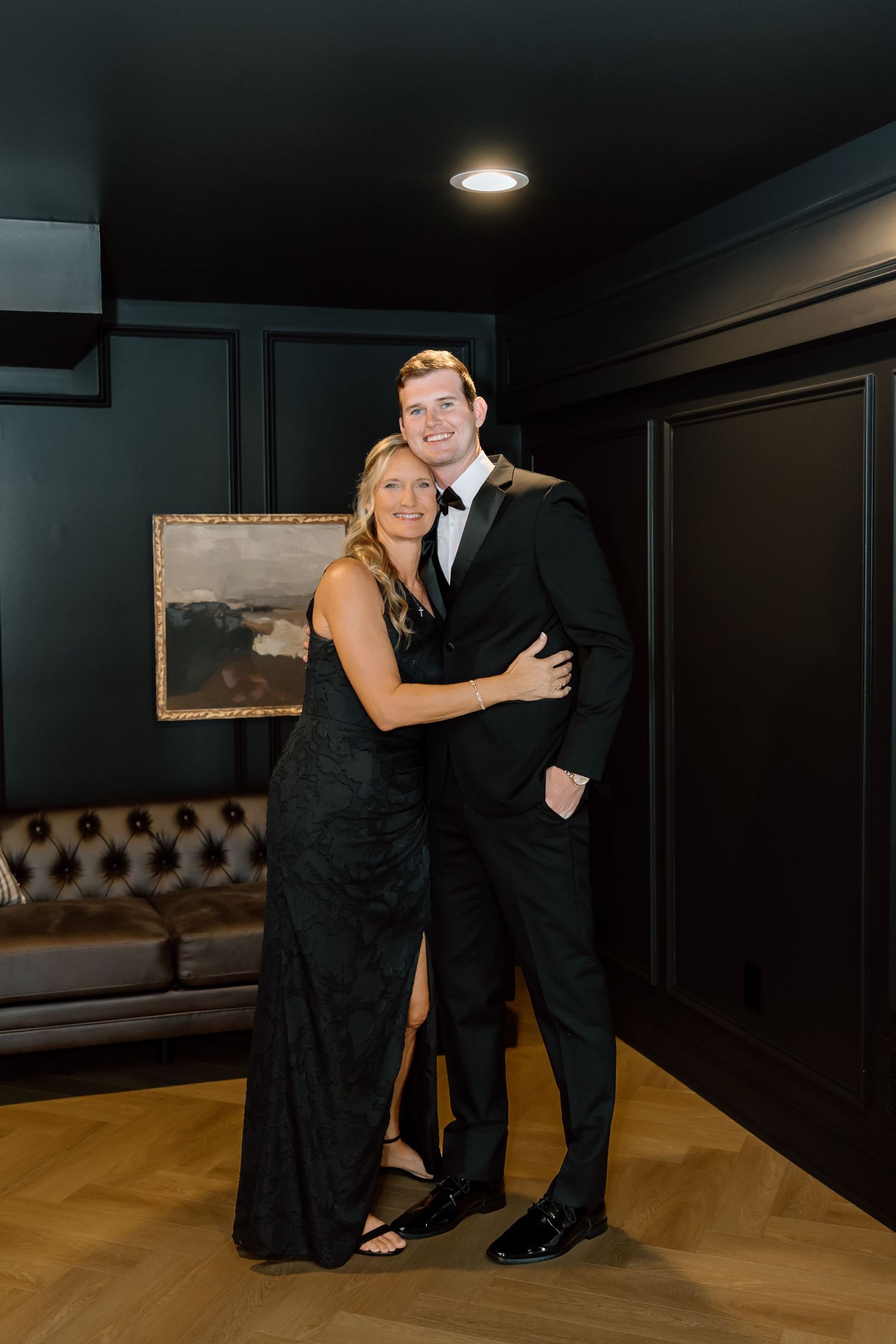 A couple in elegant black formal attire smiling and embracing in a sophisticated dark-walled venue.