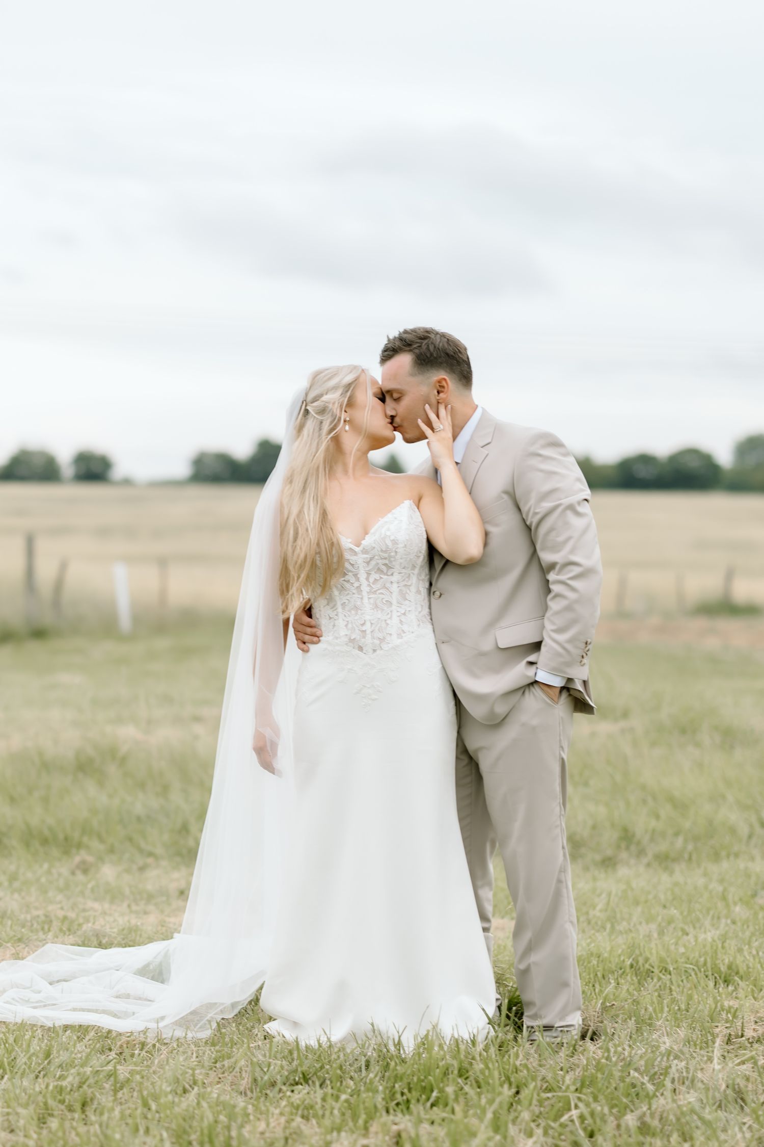 A bride and groom share a romantic kiss in a lush green field, she in a white gown with veil, he in a tan suit.