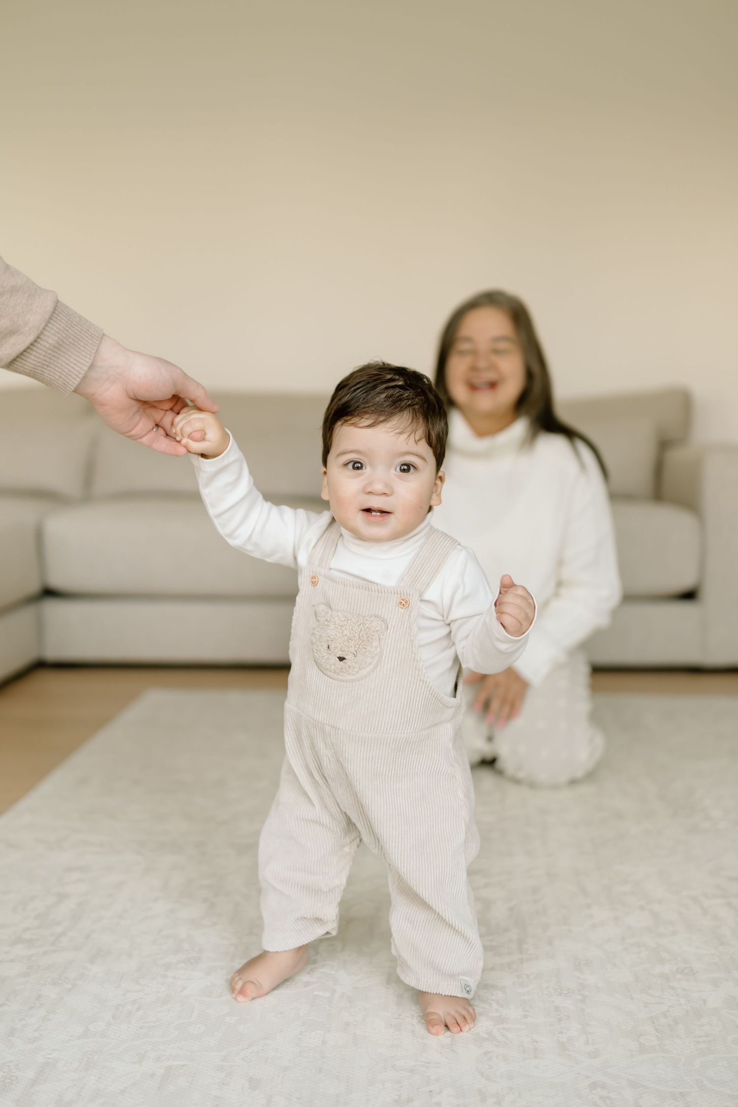 A baby in white clothing learning to walk on a light carpet while being supported by helping hands.
