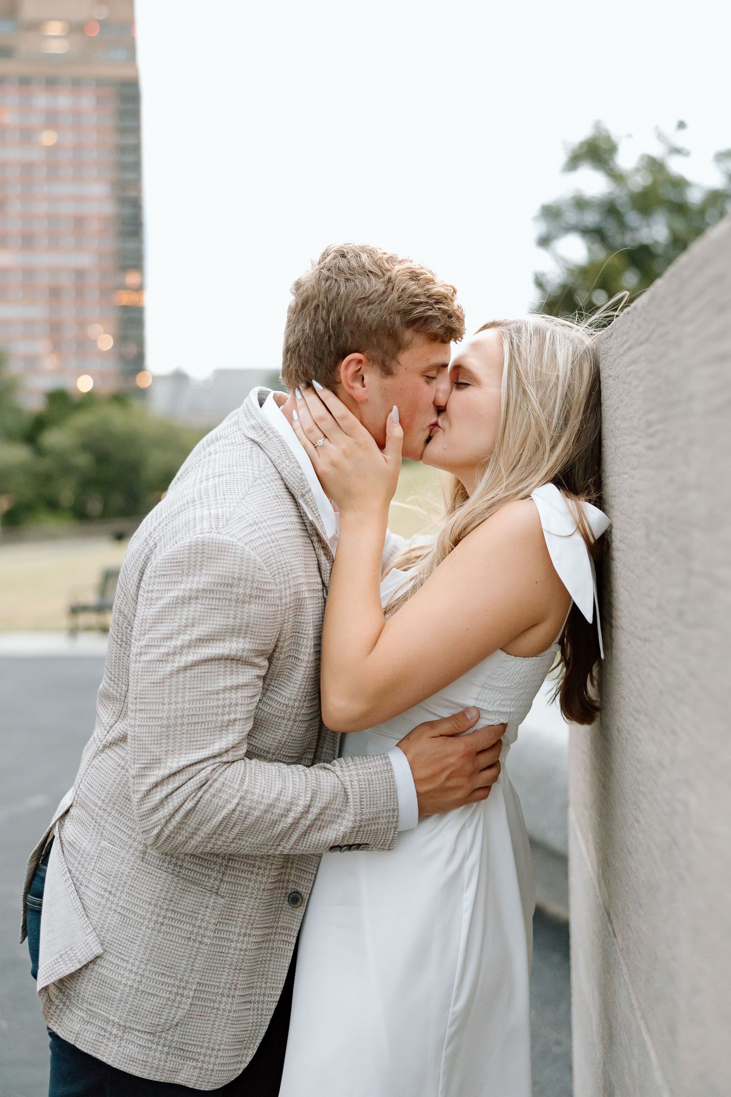 A couple shares a romantic kiss outdoors against an urban backdrop with tall buildings visible in the background.