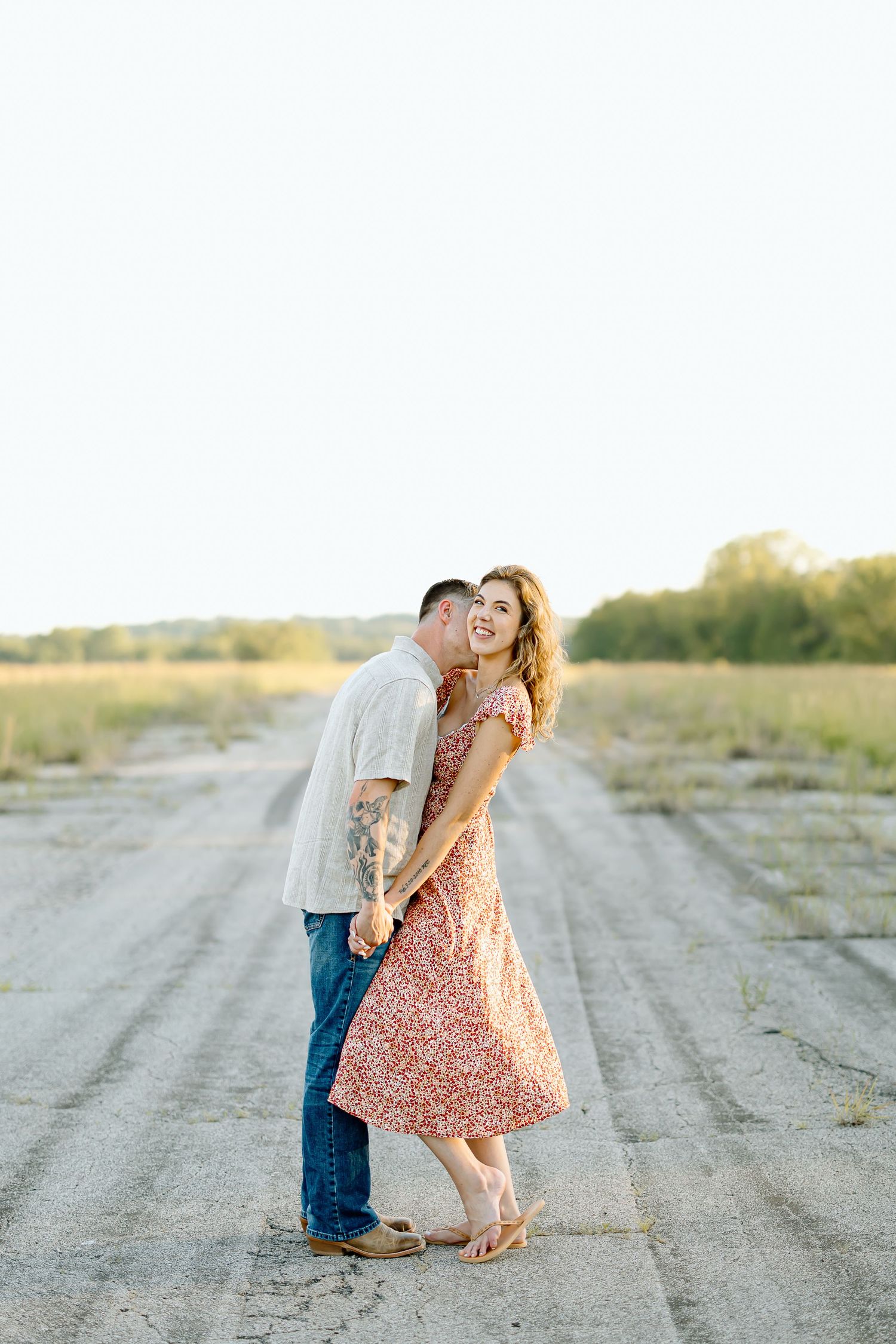 A couple shares a romantic moment on a dirt road during a sunset photoshoot in a rural setting.