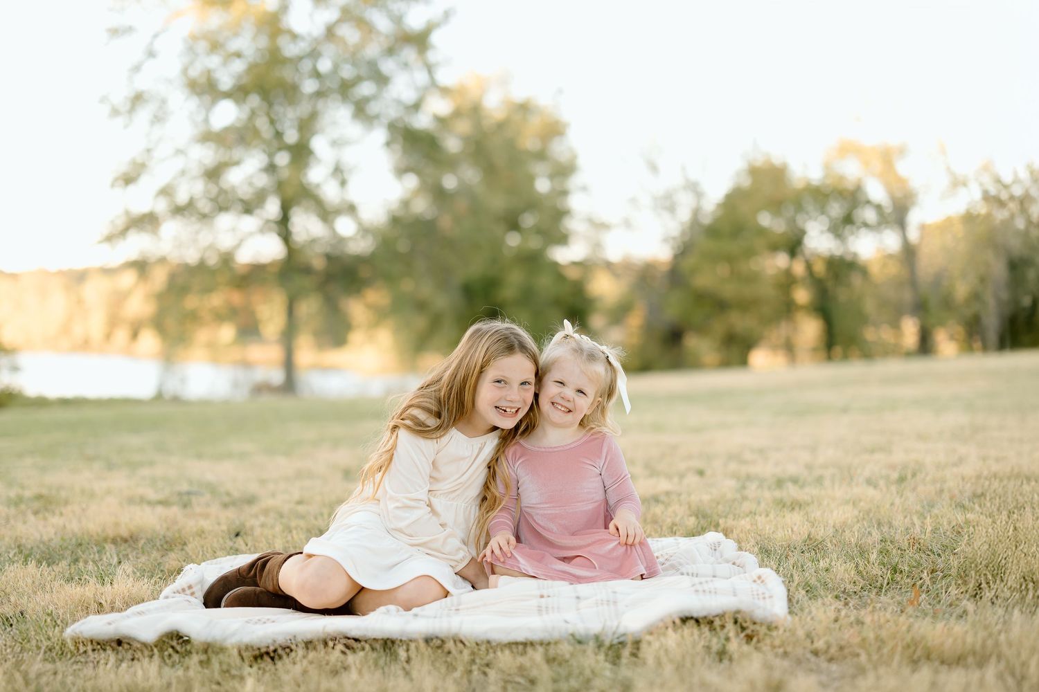 Two people sit together on a white blanket in a sunlit grassy field during golden hour.