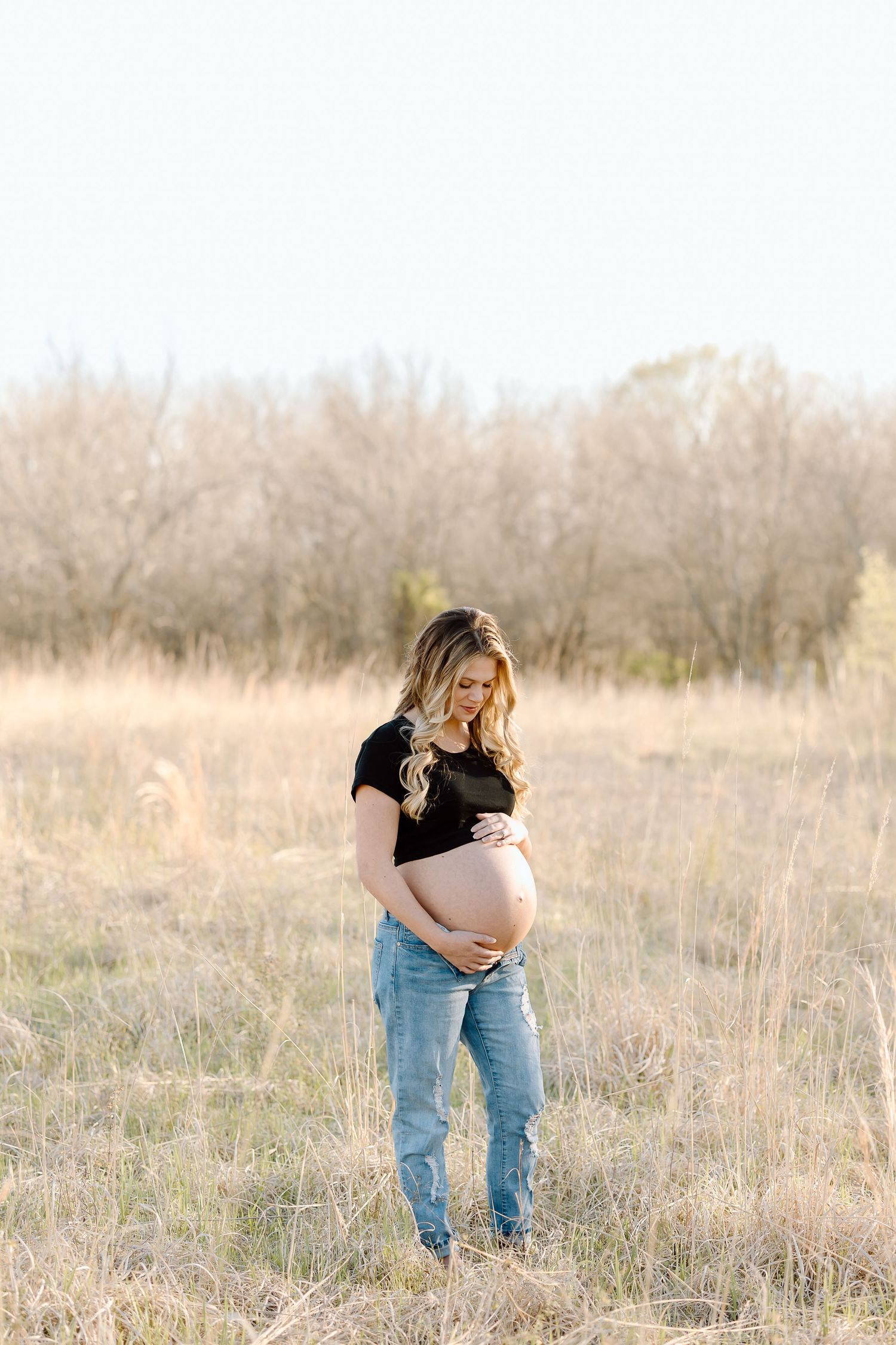 A pregnant woman in jeans and black shirt stands in a sunlit field with dried grass and bare trees in background.