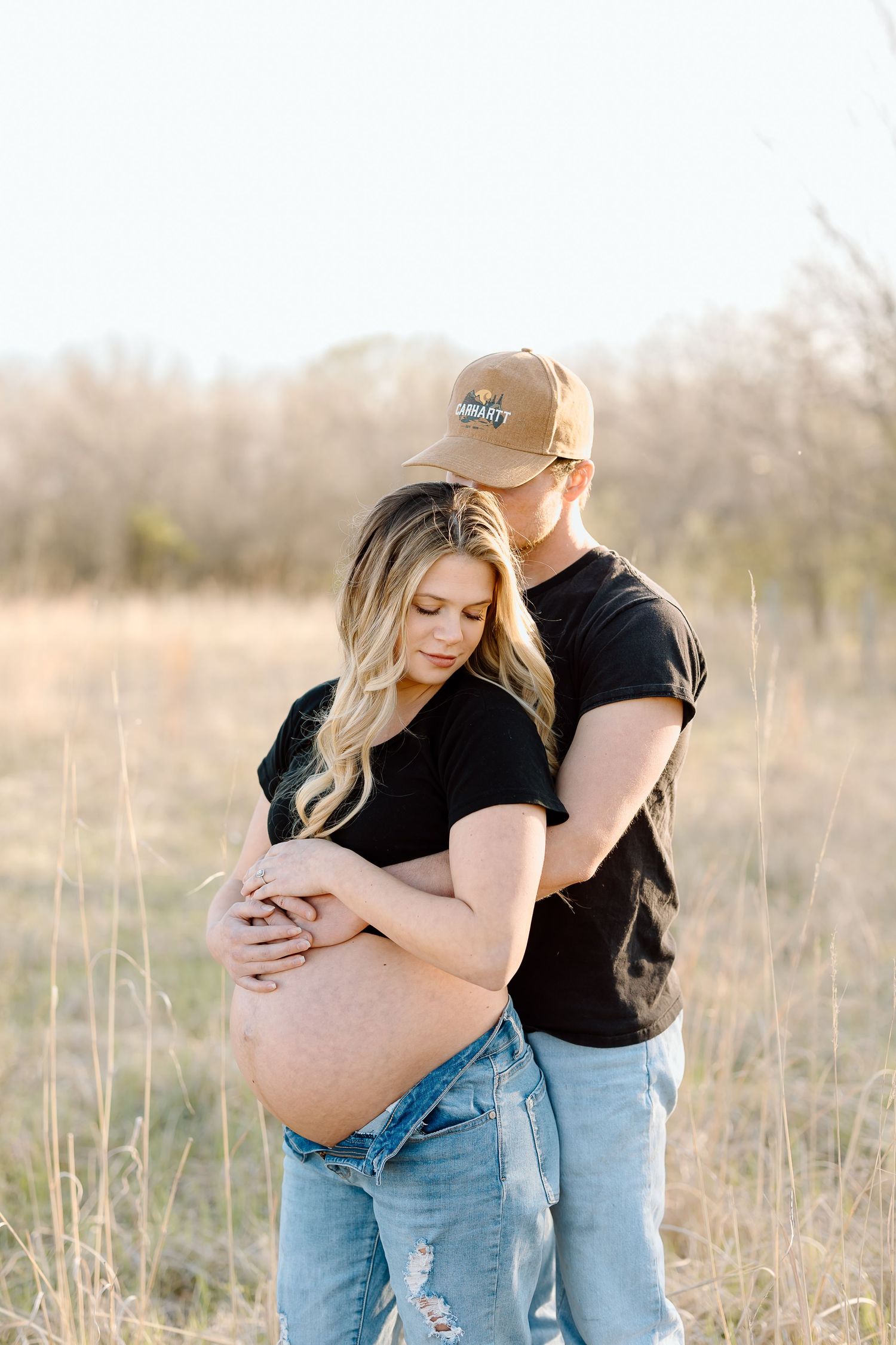 A couple embraces in a sunlit field during a maternity photoshoot wearing matching black shirts and jeans.