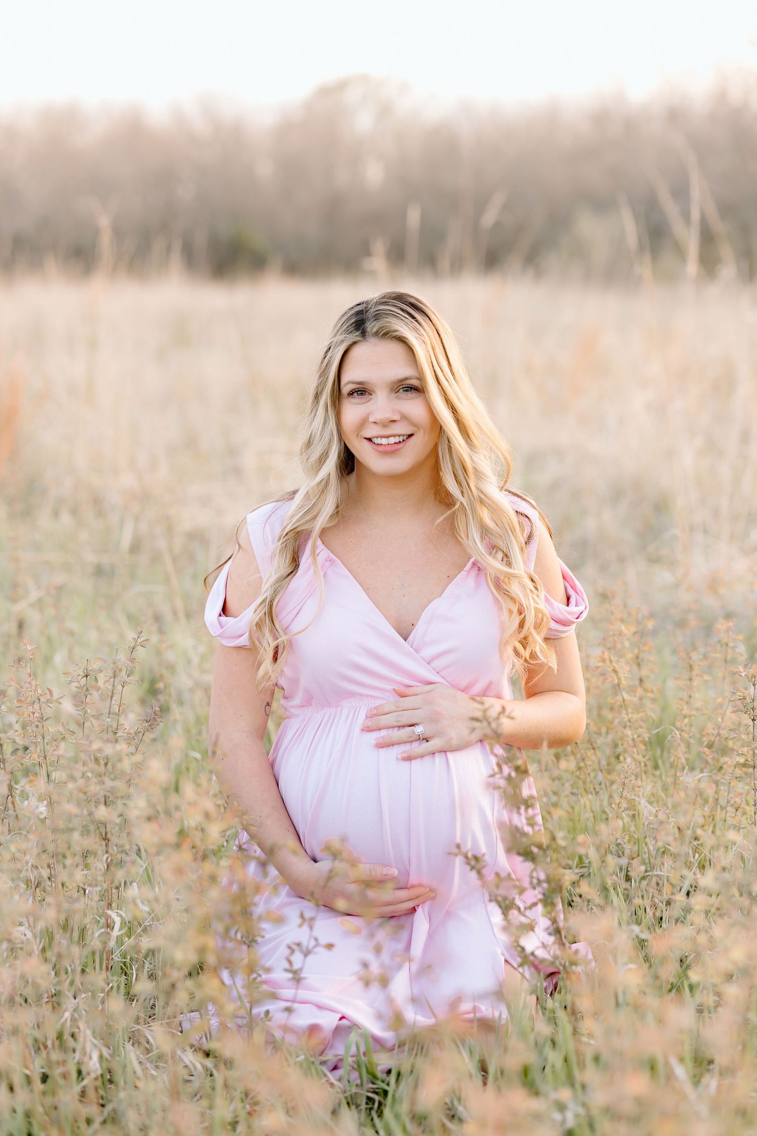 A person in a pink dress stands in a golden field during sunset, cradling their belly in a maternity photo.