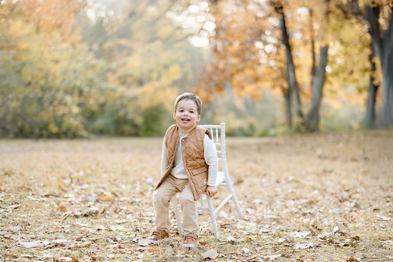 A small figure in tan clothing plays joyfully among fallen autumn leaves in a park setting.