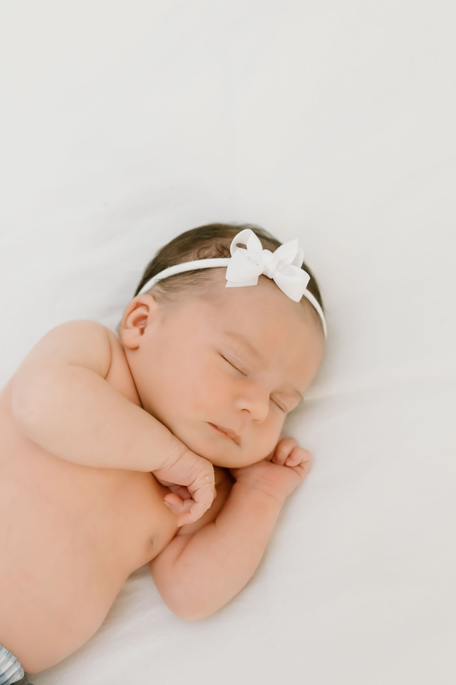 A sleeping newborn baby in white swaddling cloth with a small bow headband rests peacefully against a light background.