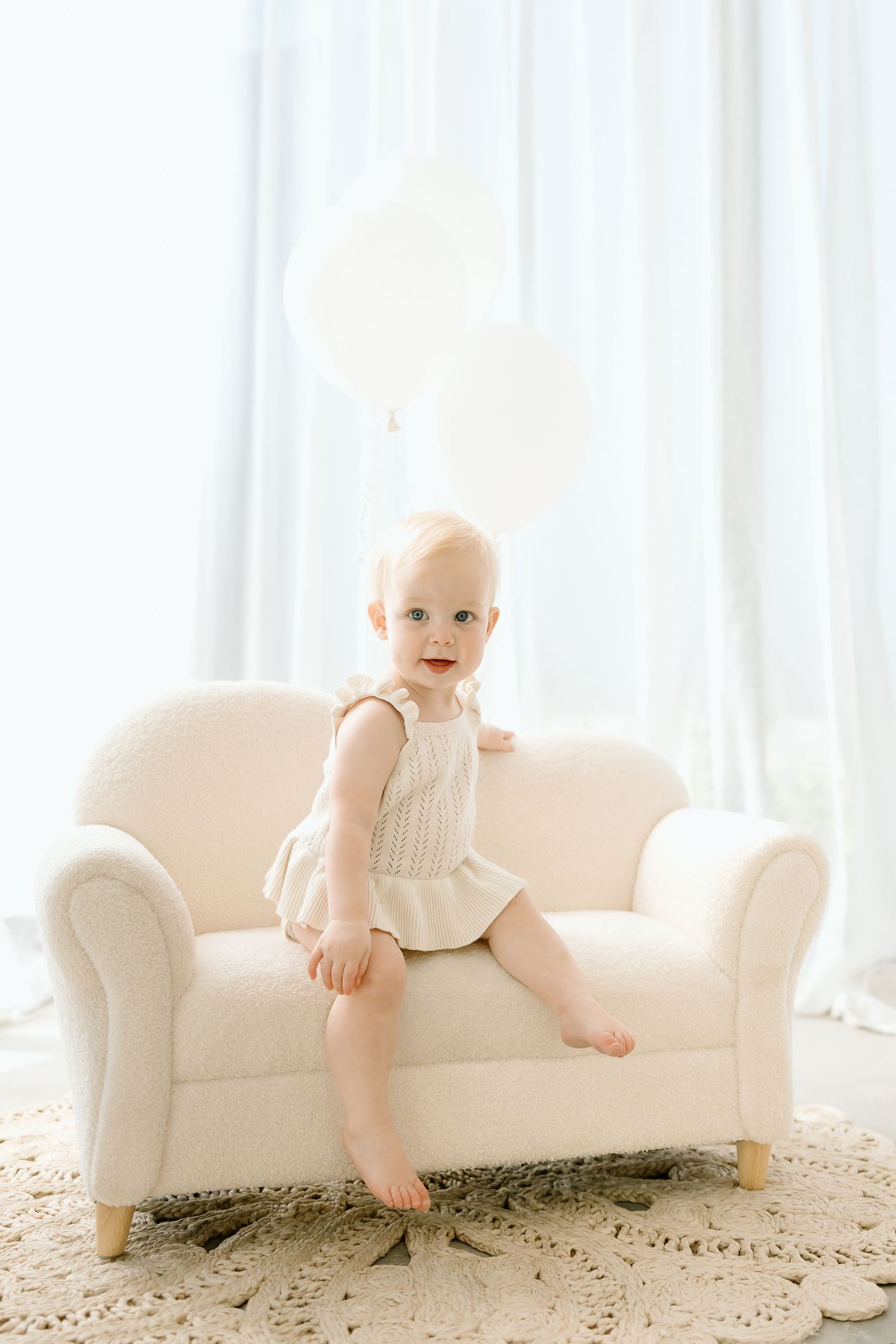 Baby in white outfit sits on cream-colored couch in bright, airy room with soft window light.