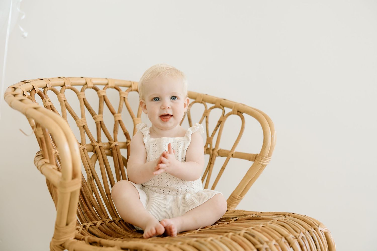 Baby sitting in a woven rattan peacock chair wearing white against a neutral background.