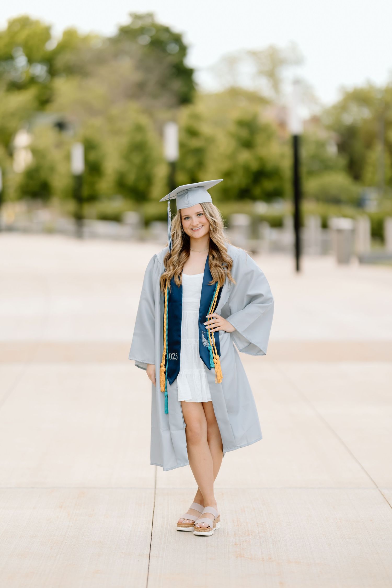 Graduate in blue and gray cap and gown poses outdoors for graduation photos on a sunny day.