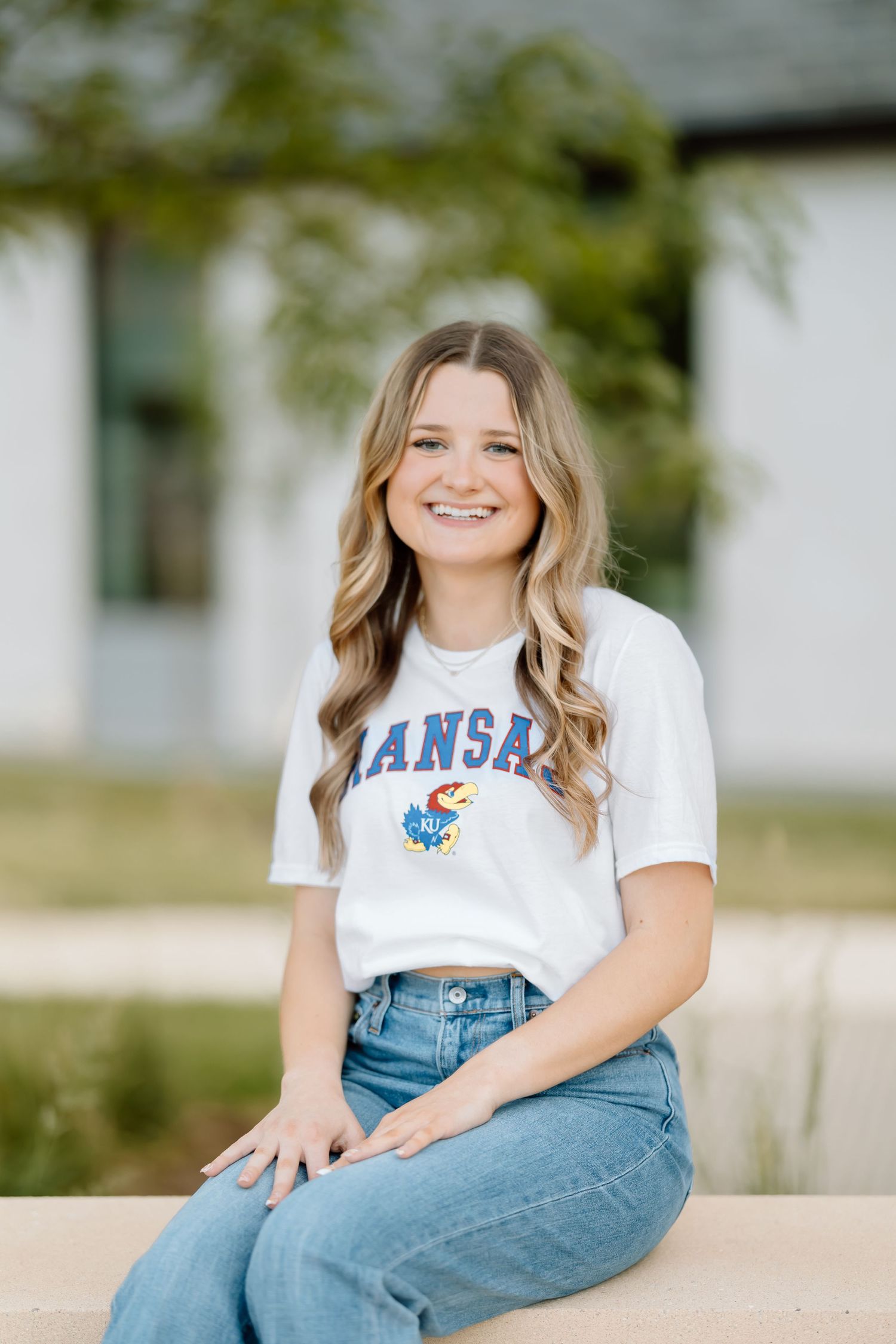 Student wearing a Kansas University t-shirt and jeans sits casually outdoors on a sunny day.