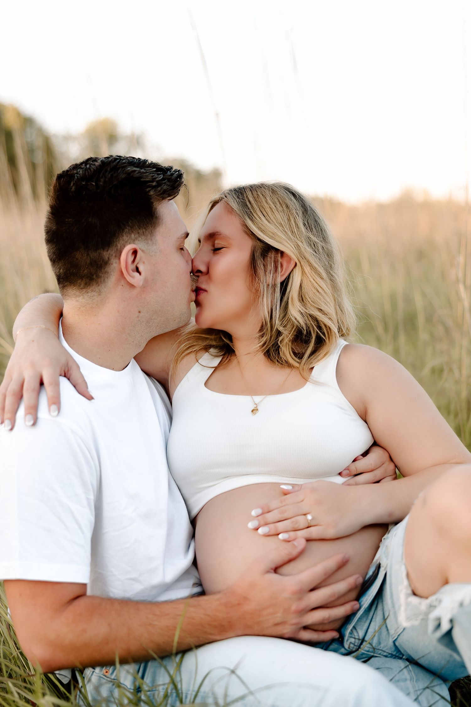 A romantic couple embraces and kisses in a sunlit field of tall grass during a golden hour photoshoot.