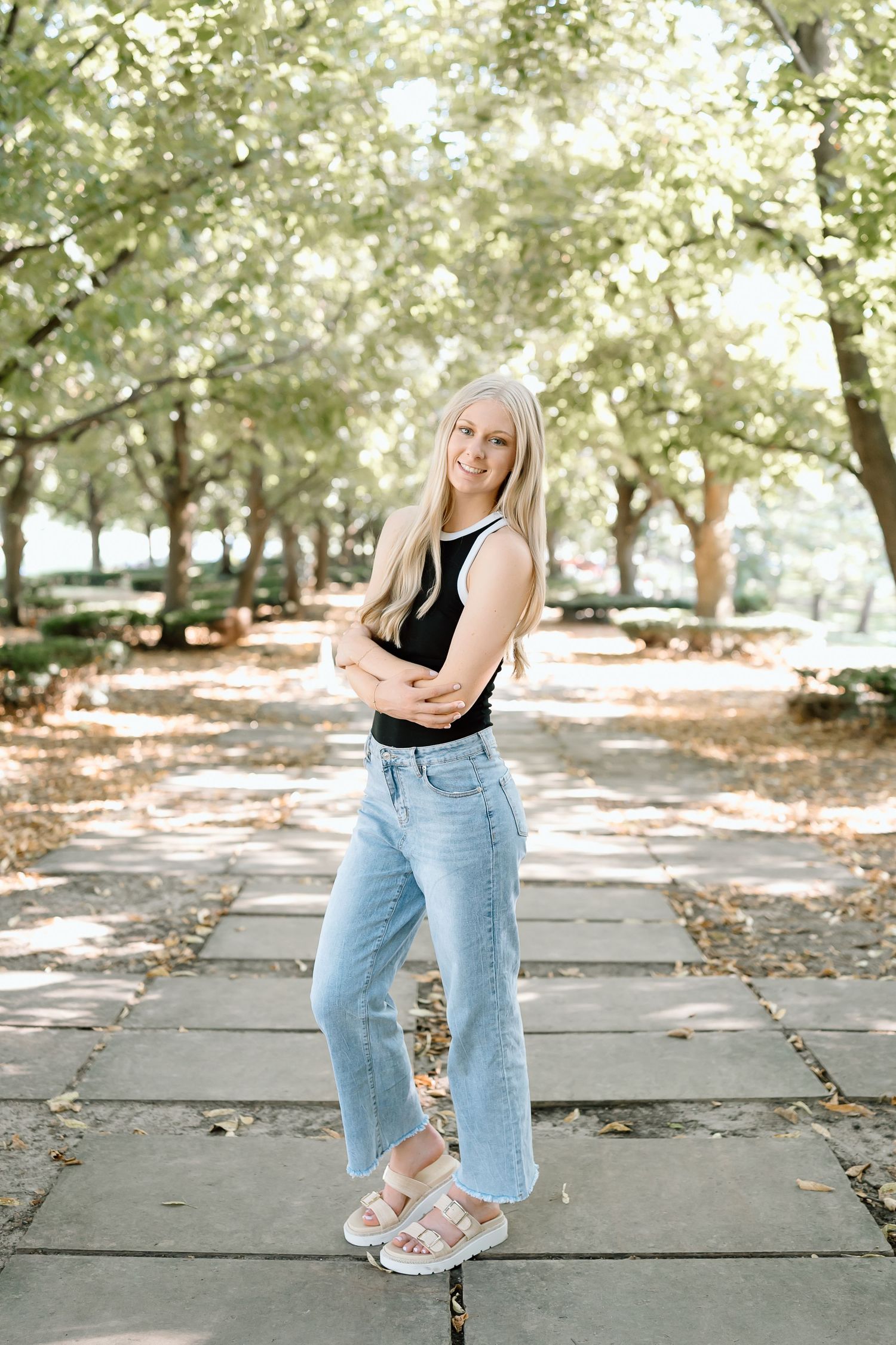 A person in a black tank top and light wash jeans stands on a stone path surrounded by sunlit trees in a park.
