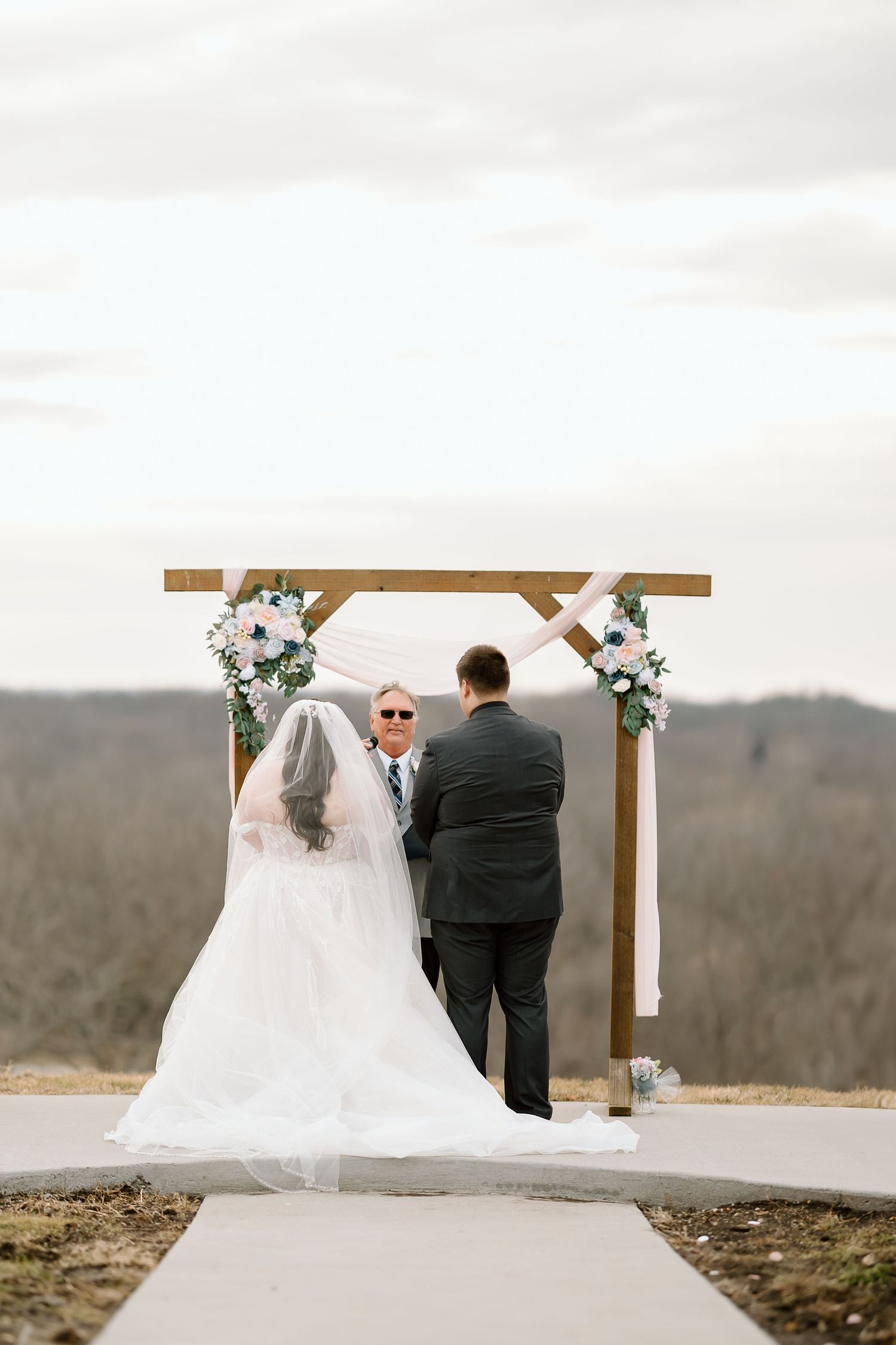 An outdoor wedding ceremony takes place under a wooden arch decorated with white flowers on a hilltop overlook.
