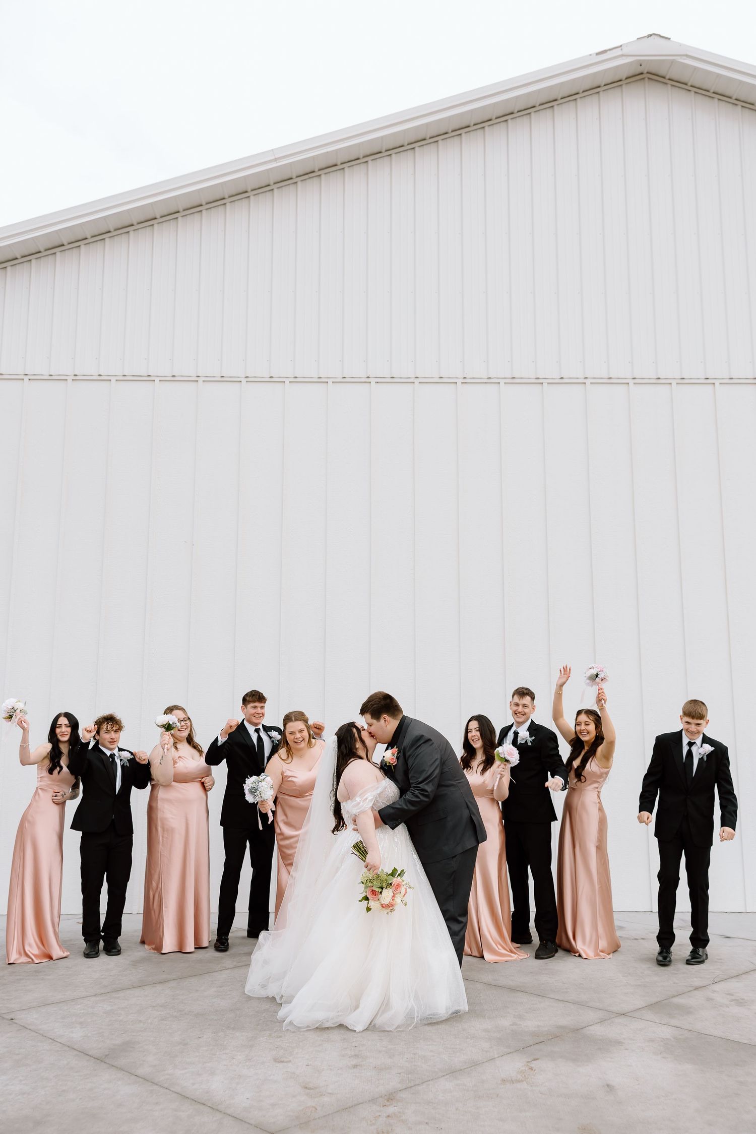 Wedding party in black and blush pink attire poses in a fun leaning formation against a white wall.