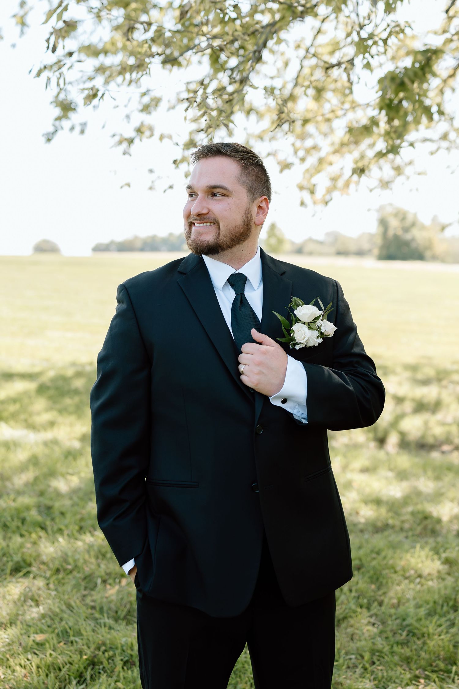 A groom in a black tuxedo stands outdoors on a grassy hillside with sunlit trees in the background.