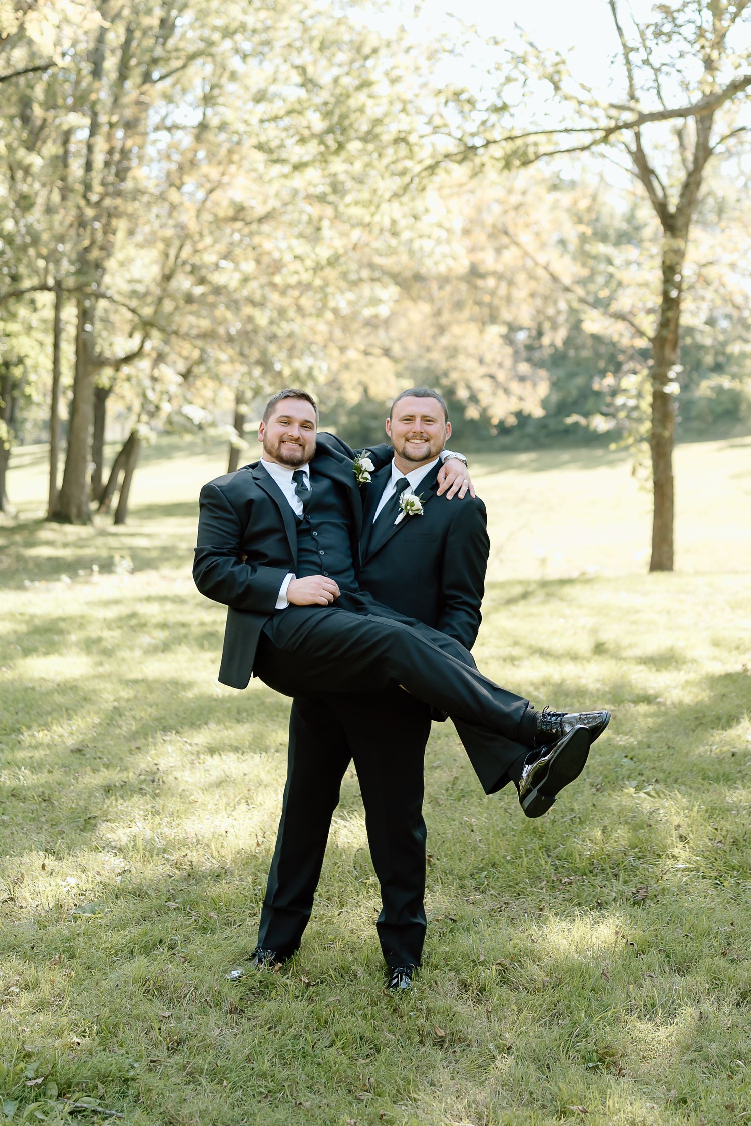 Two people in black tuxedos share a playful moment outdoors on their wedding day in a sunlit wooded setting.