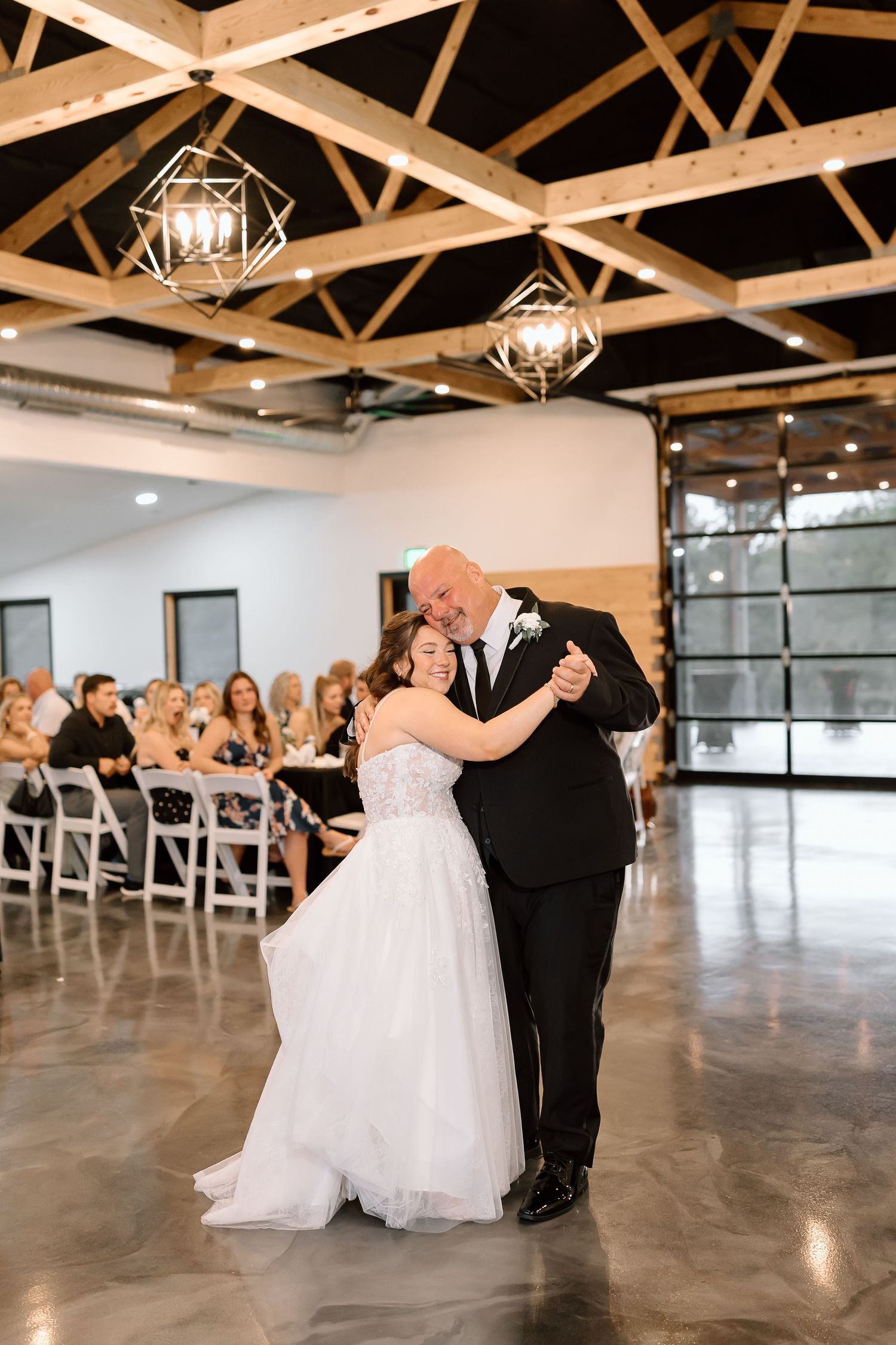 A couple shares their first dance at a modern reception venue with exposed wooden beams and geometric light fixtures.