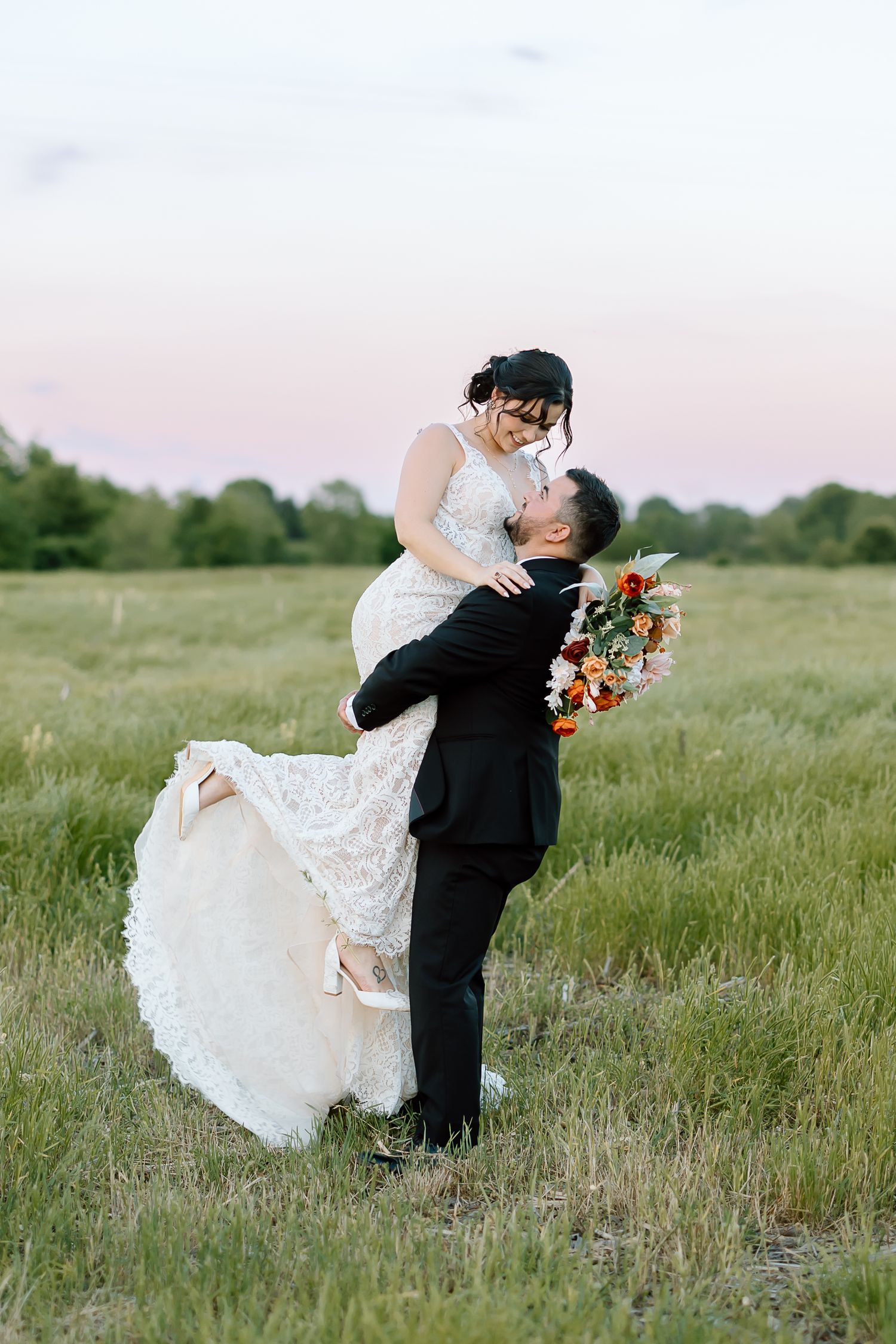 A couple in wedding attire shares a romantic embrace while dancing outdoors in a scenic meadow at sunset.