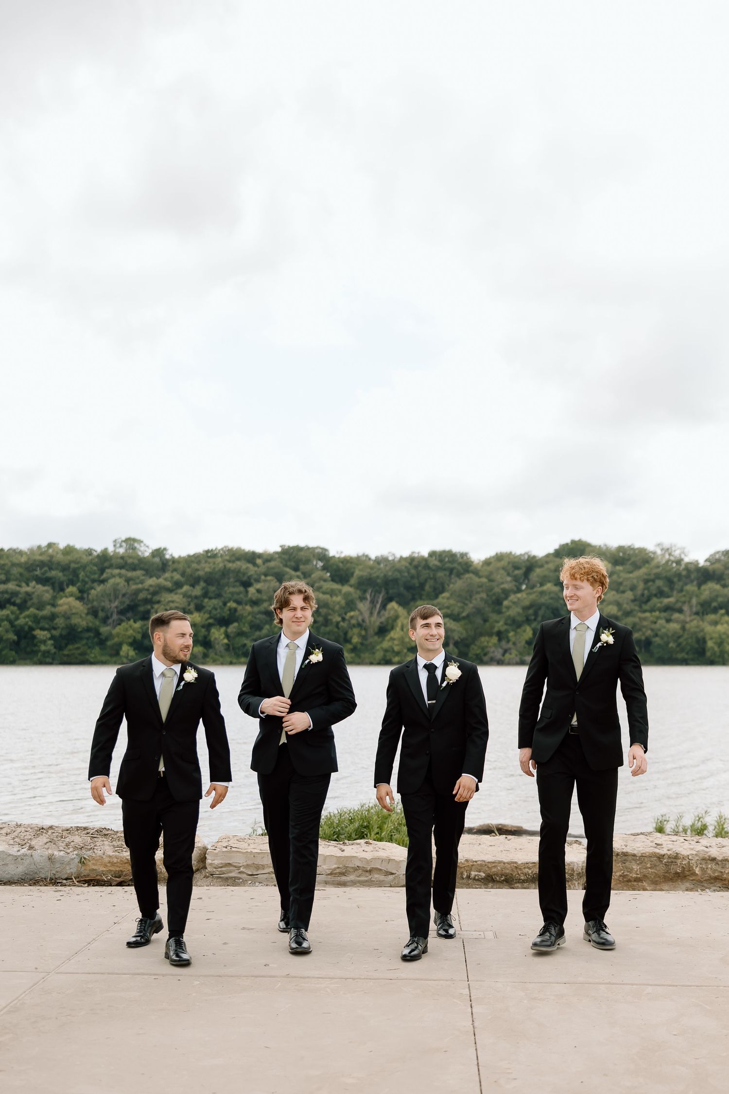 Four groomsmen in black suits and boutonnieres stroll along a lakeside path on a cloudy day.