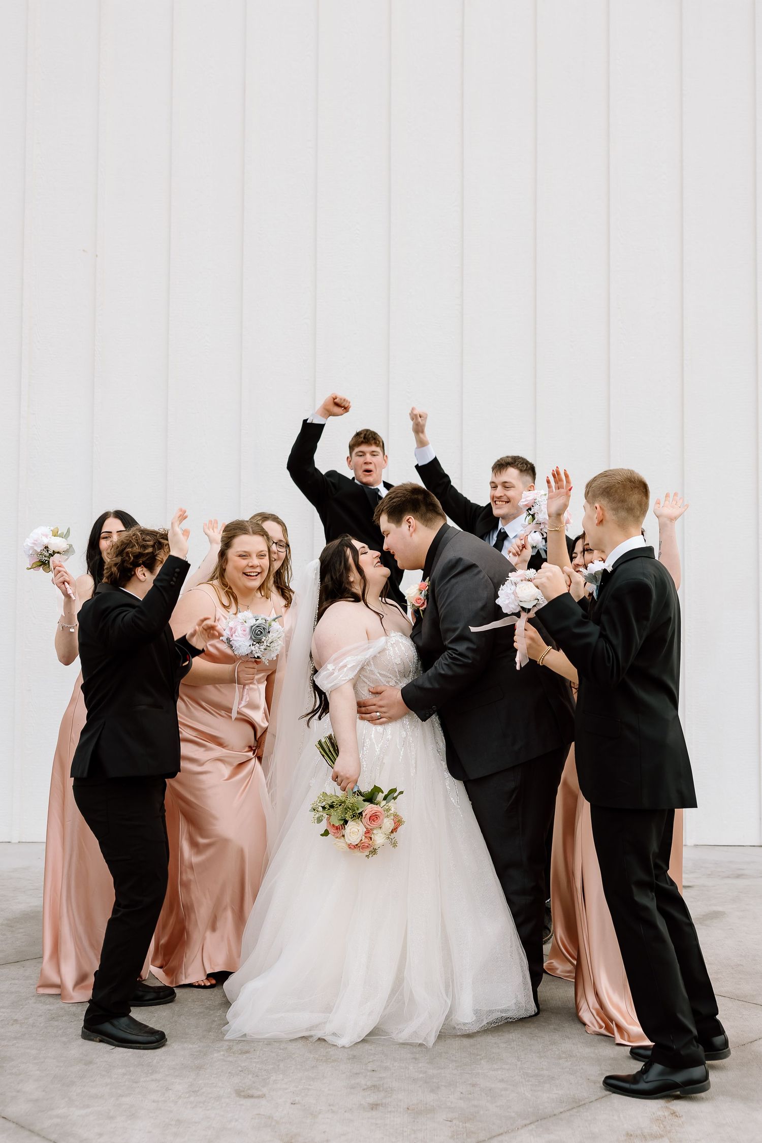 Wedding party celebrates enthusiastically against white building backdrop.