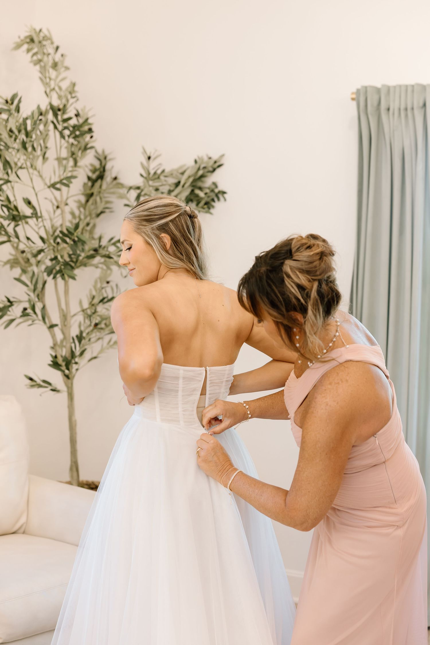 Helper assisting with wedding dress buttons in a light-filled room with greenery decor.
