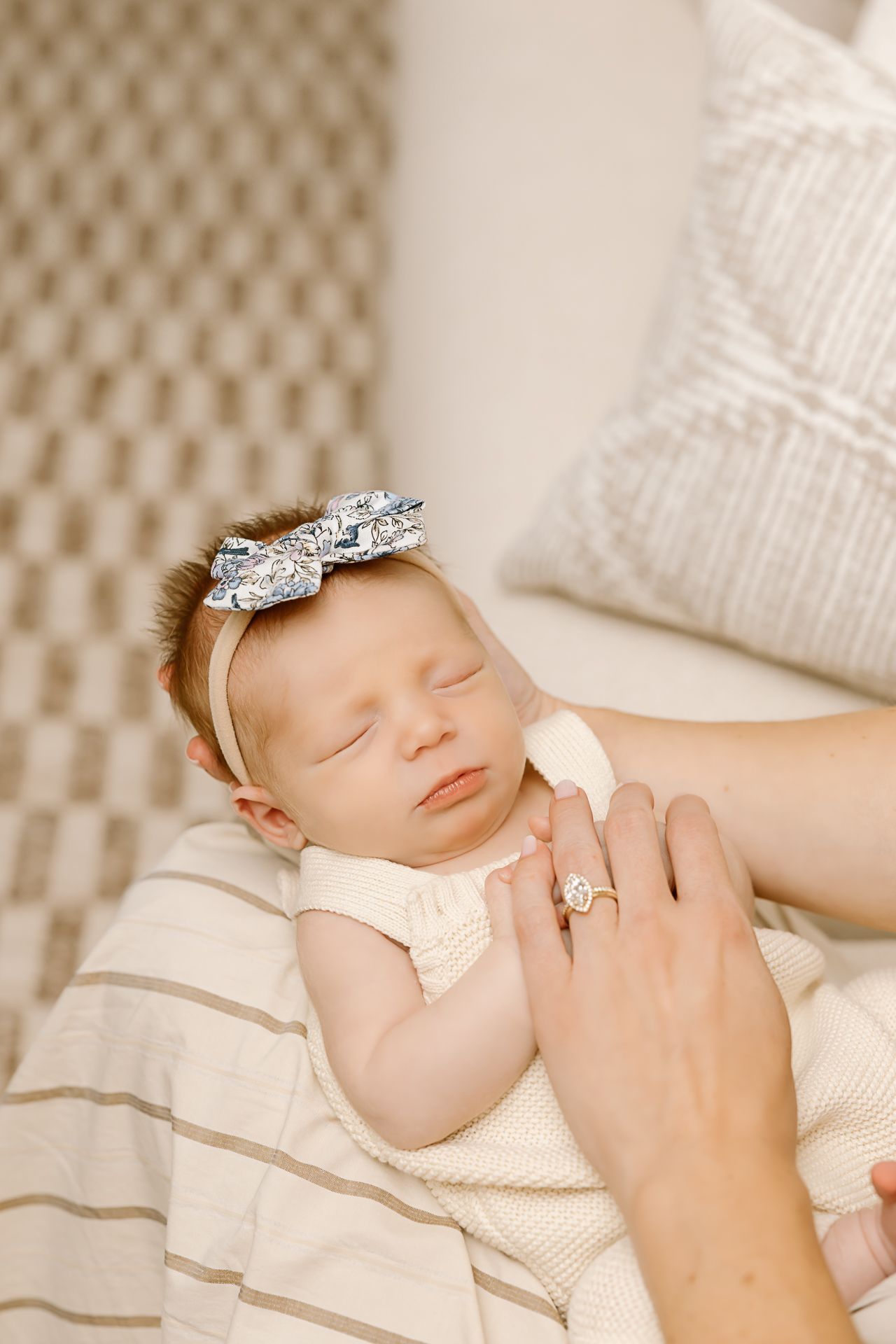 Sleeping baby in cream outfit with sparkly bow headband and parent's hand showing ring.