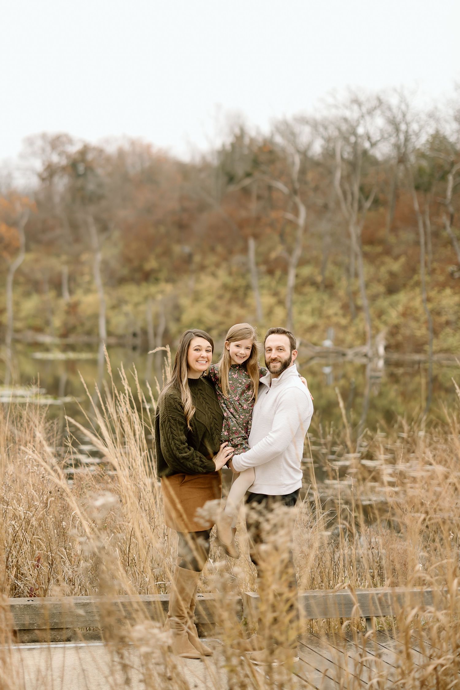 Family standing among tall grass near autumn lake with trees in background.