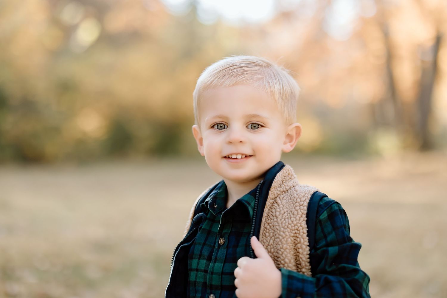 Young child in plaid shirt and sherpa vest smiles outdoors during fall season.