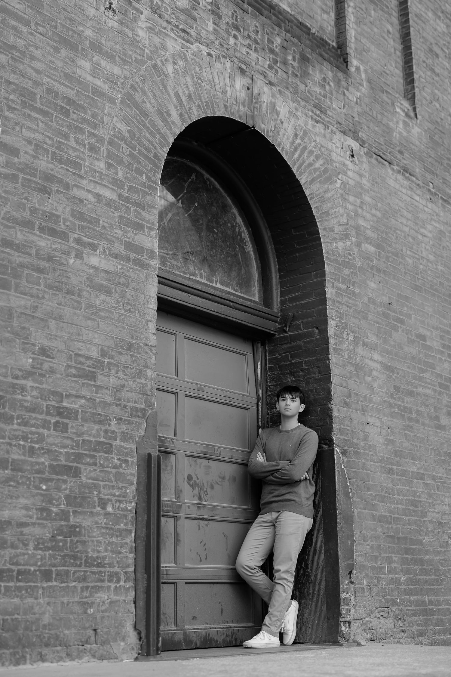 Black and white portrait of person leaning in arched doorway of brick building.