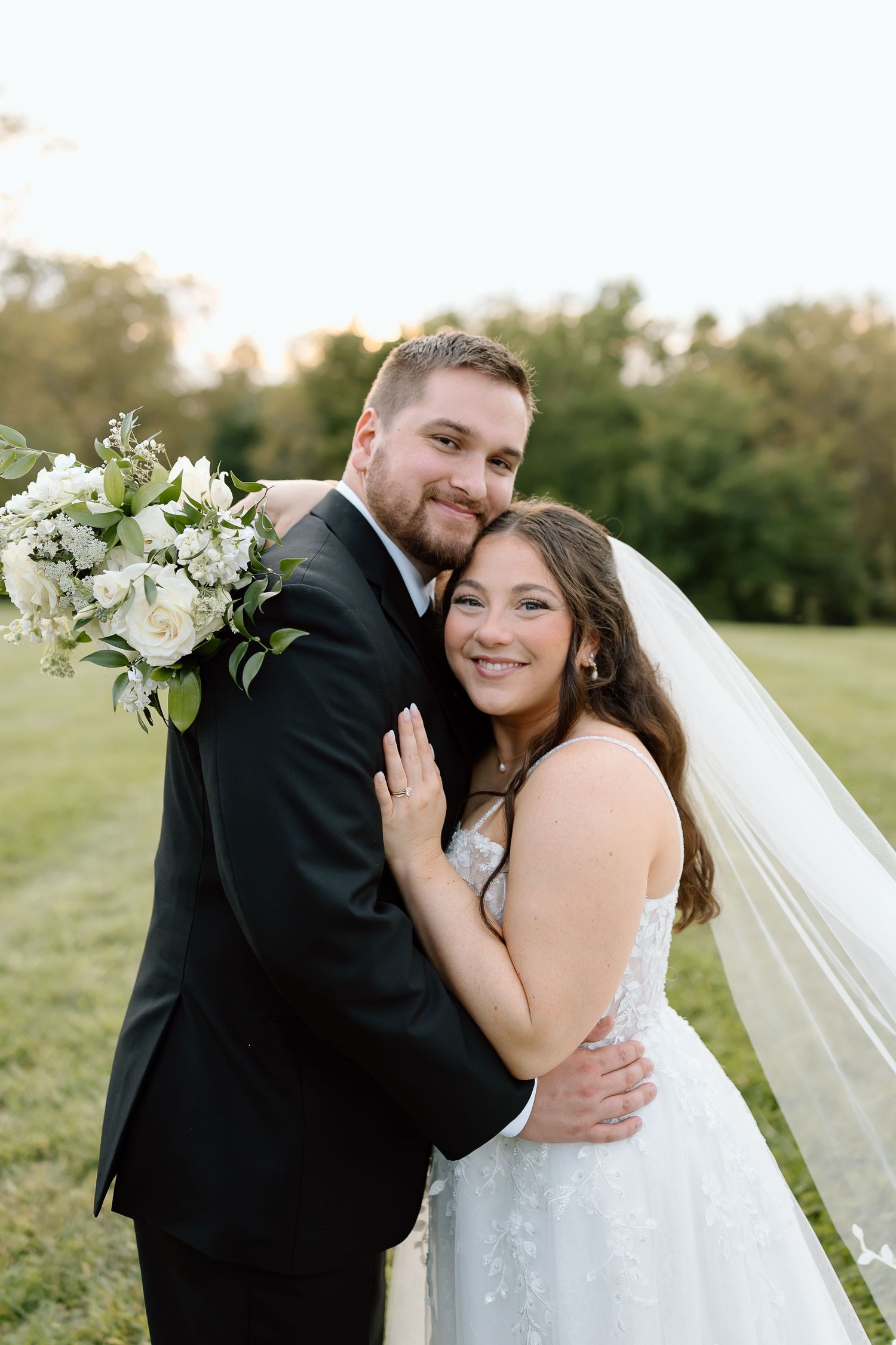 A romantic wedding couple embraces outdoors in a grassy field at sunset.