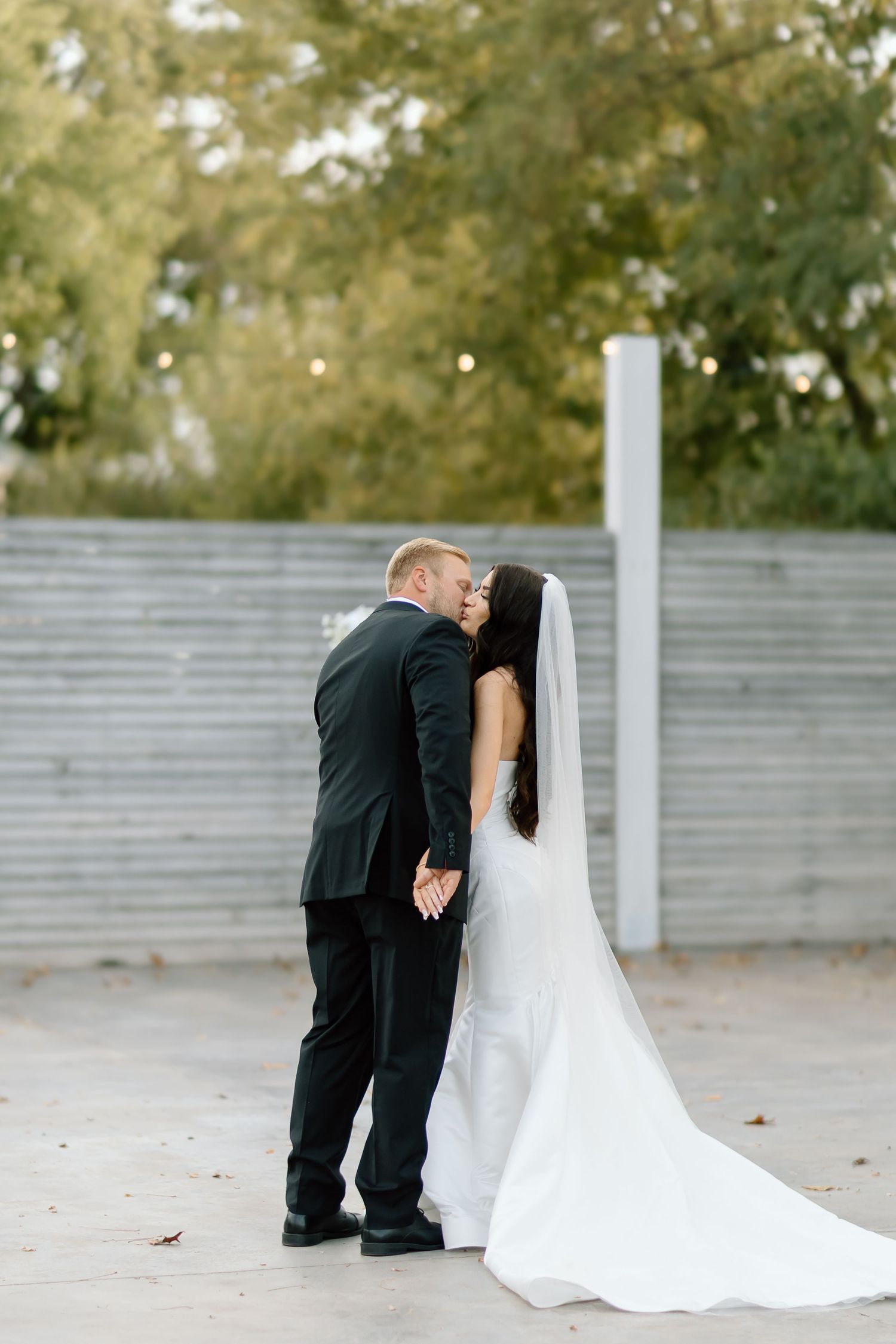 A romantic wedding couple embraces in a tender moment against a soft outdoor backdrop with white fencing.