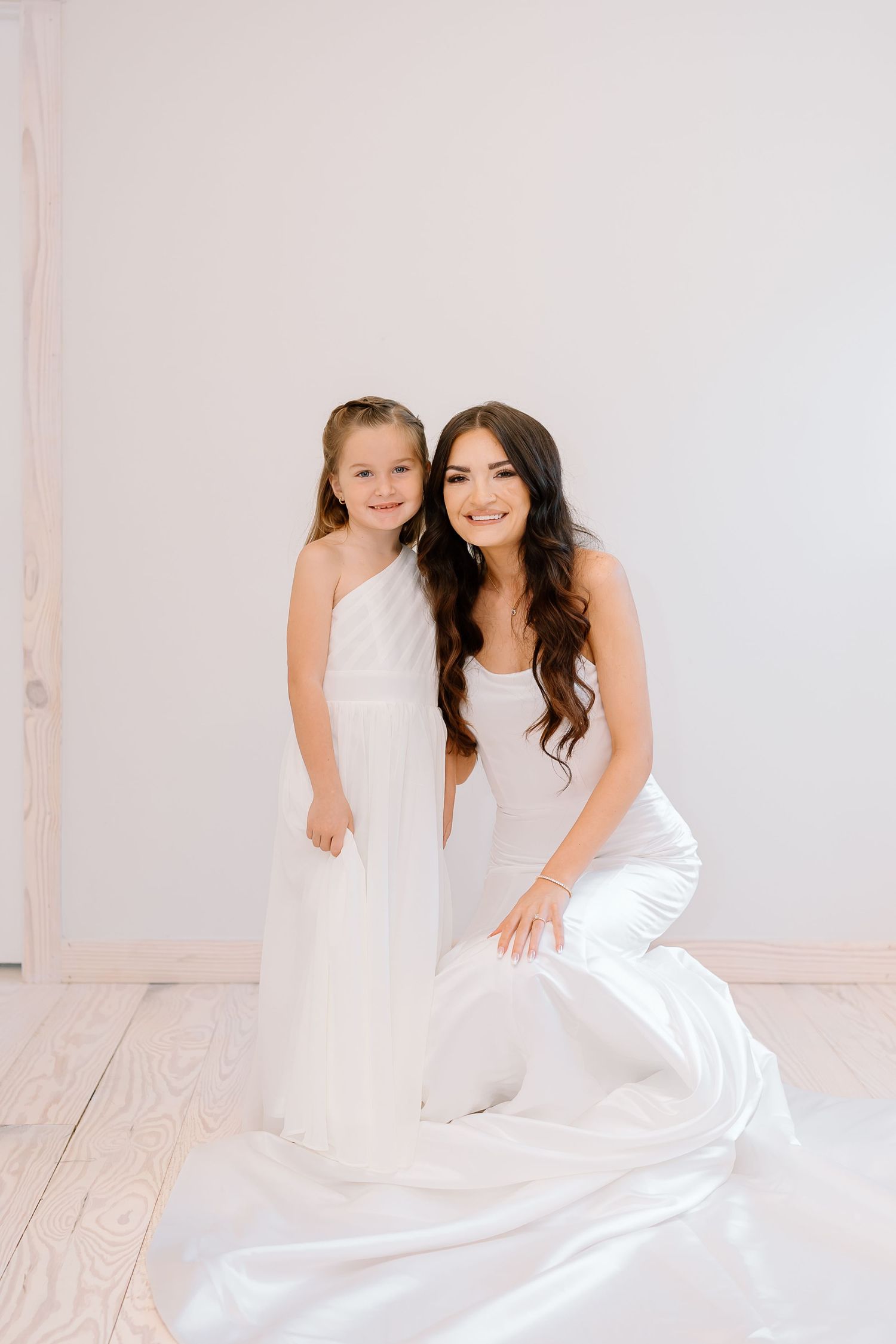 Two people in elegant white dresses pose together in a bright minimalist setting with light wooden floors.