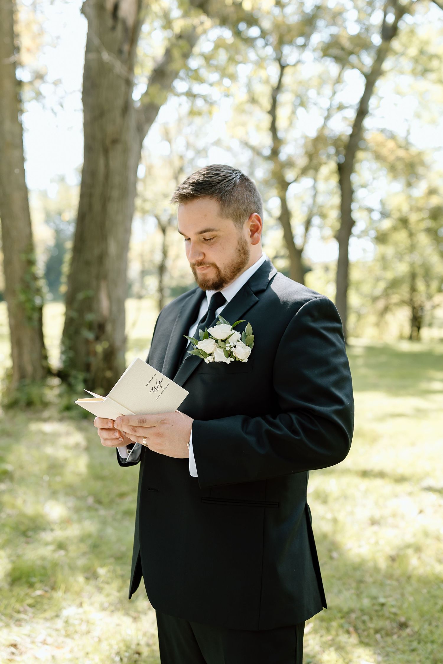 A groom in a black suit reads a letter outdoors in a sunlit wooded area during a wedding day.