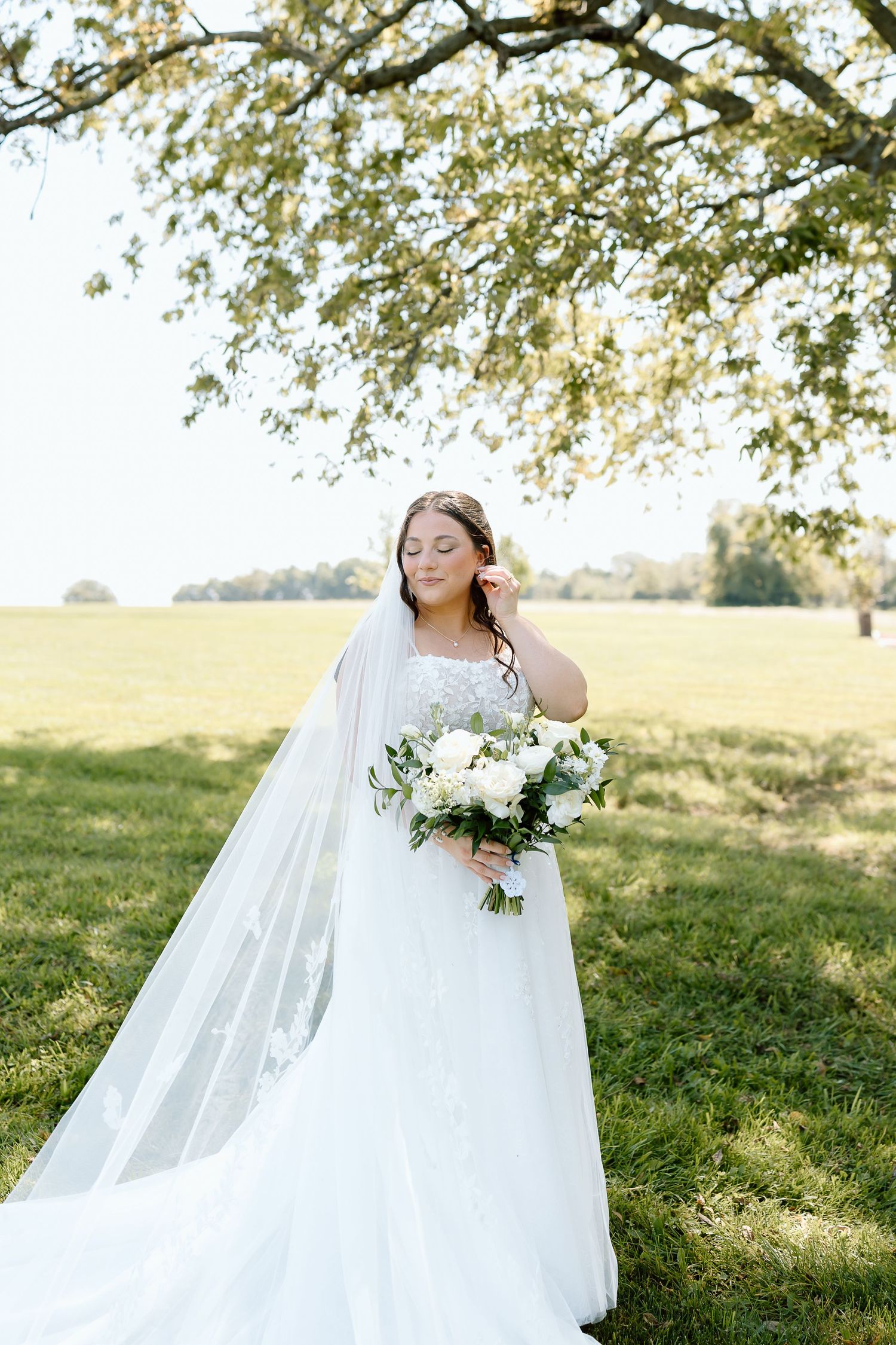 A bride in a flowing white wedding dress twirls joyfully in a sunlit meadow with trees in the background.