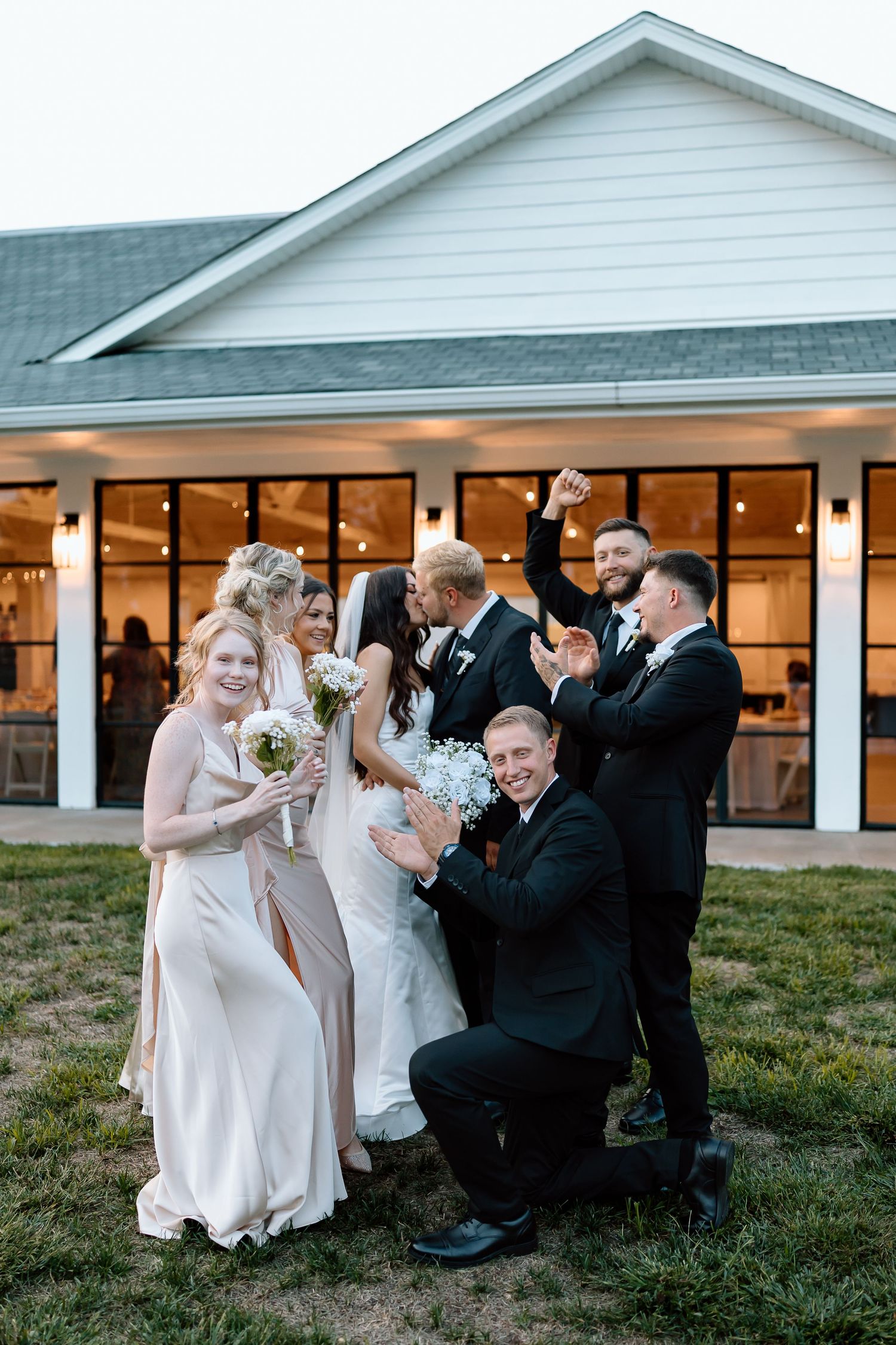 Wedding party in formal attire poses playfully on grassy lawn at sunset outside venue.