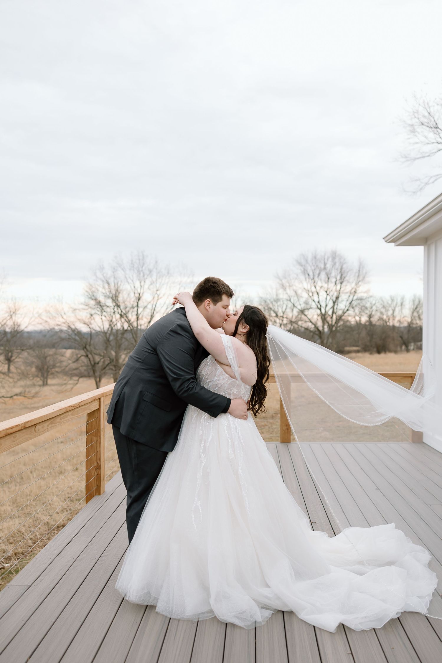 Romantic wedding couple shares a kiss on a rustic wooden deck overlooking a scenic winter landscape.
