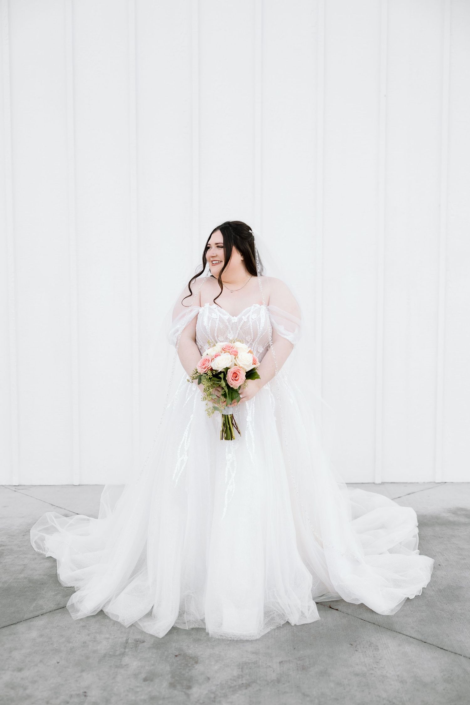 A bride in a flowing white wedding gown with pink sleeves holds a pink and white floral bouquet against a light backdrop.