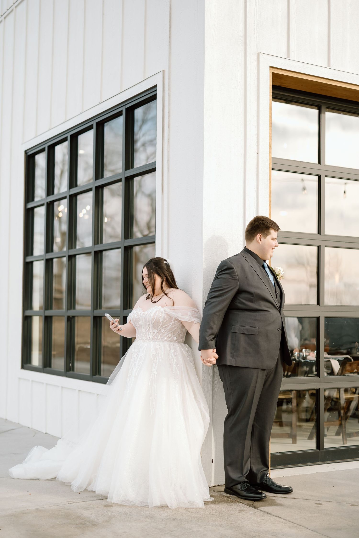 A bride and groom share a playful moment in front of a white building with large windows.