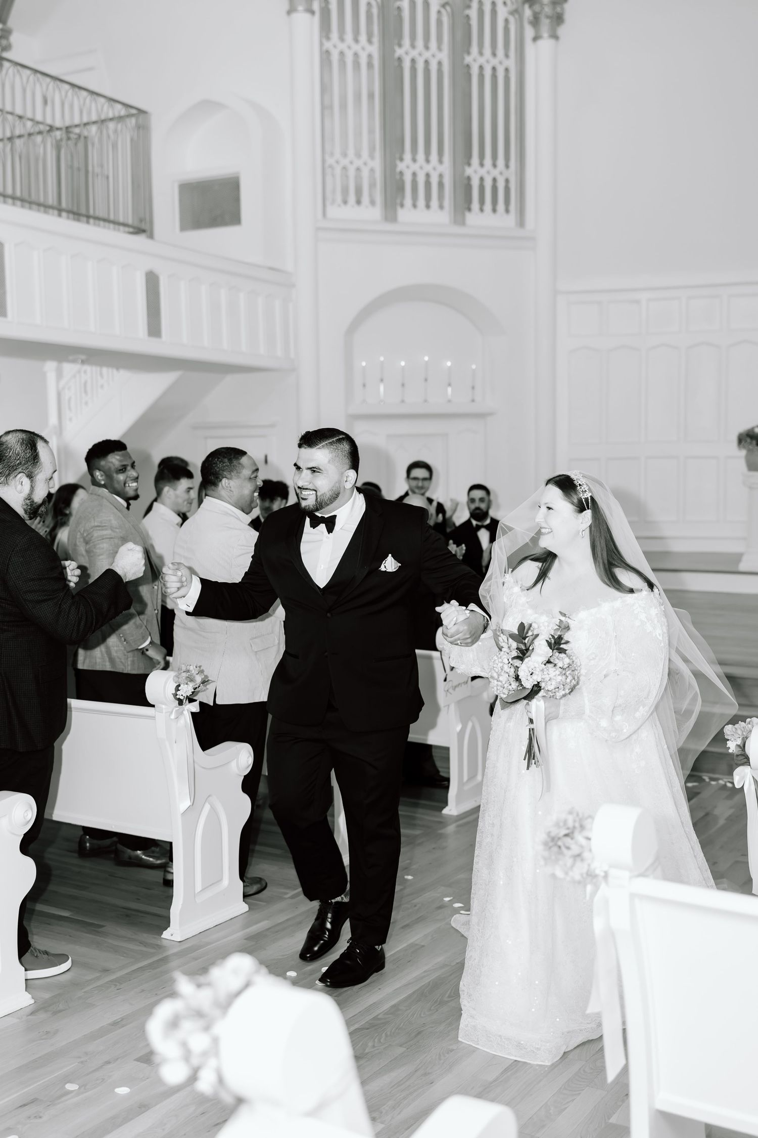Black and white photo of bride and groom walking down church aisle after wedding ceremony with guests applauding.